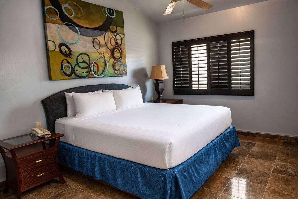 A neatly made bed with a blue skirt, a bedside lamp, a telephone, and a wall painting.