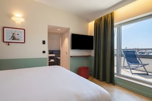 A hotel room with a bed, a TV mounted on the wall, a window with a view of the city, and a balcony with a chair. - Urban Hive Paris (Paris)