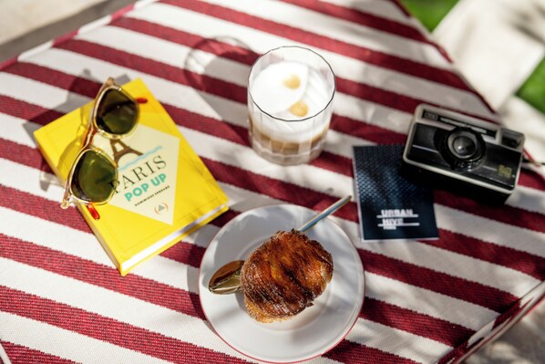 Superficie de tela a rayas con un libro, gafas de sol, una cámara, un postre y una bebida.