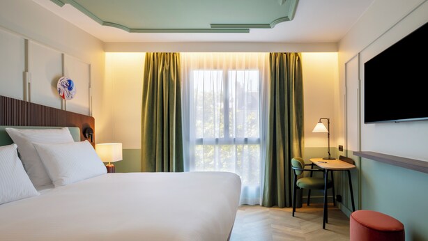 A hotel room with a large bed, a desk with a chair, a television, and a window with curtains. - Urban Hive Paris (Paris)