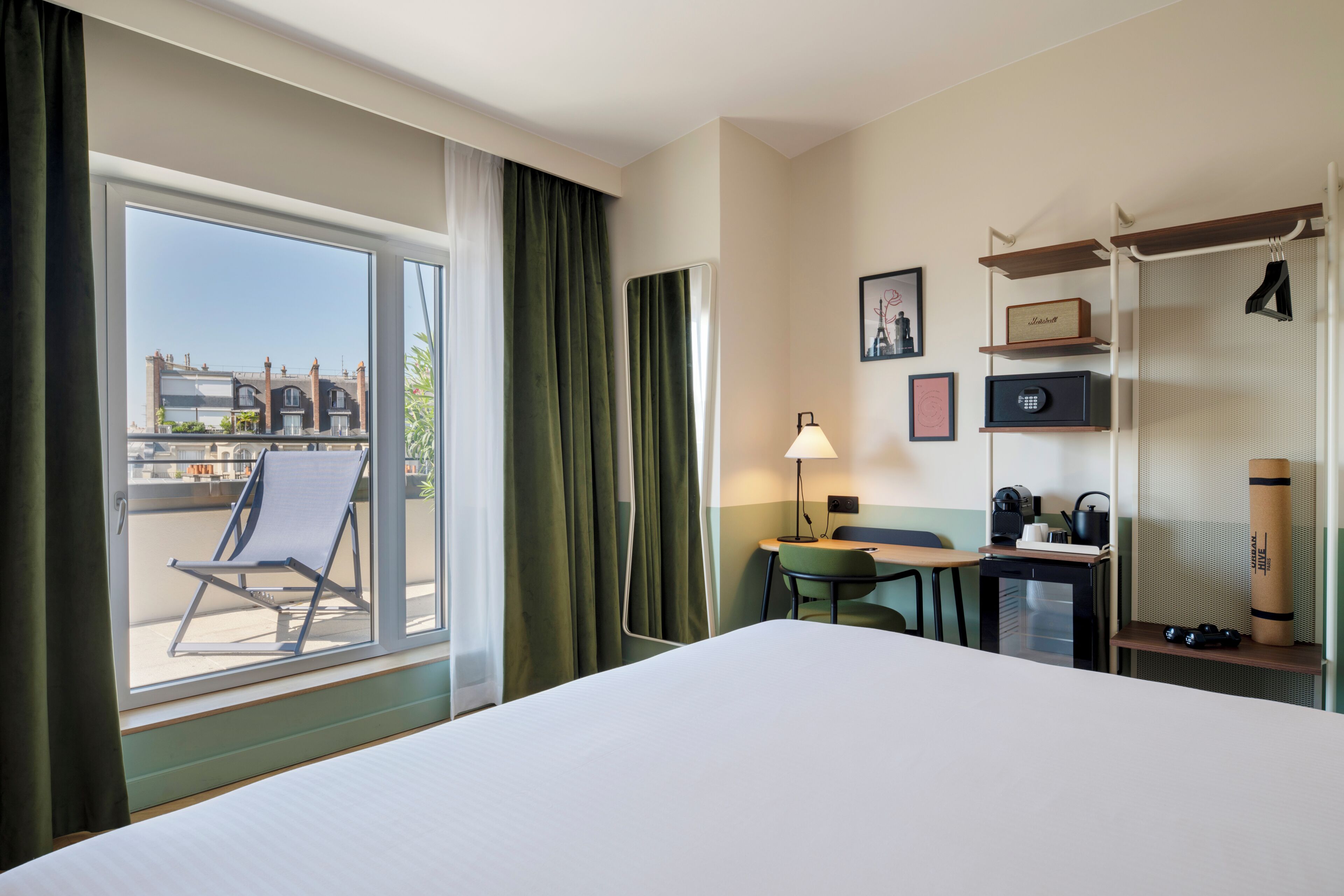 A hotel room with a bed, a desk, a chair, a lamp, a small table, a shelf with items, a window with curtains, and a view of buildings outside.