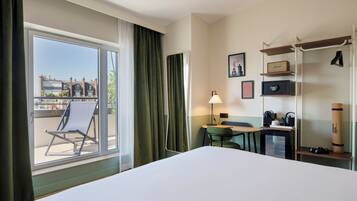 Habitación de hotel con cama, escritorio, silla, lámpara, mesita, estante con objetos, ventana con cortinas y vista a edificios.
