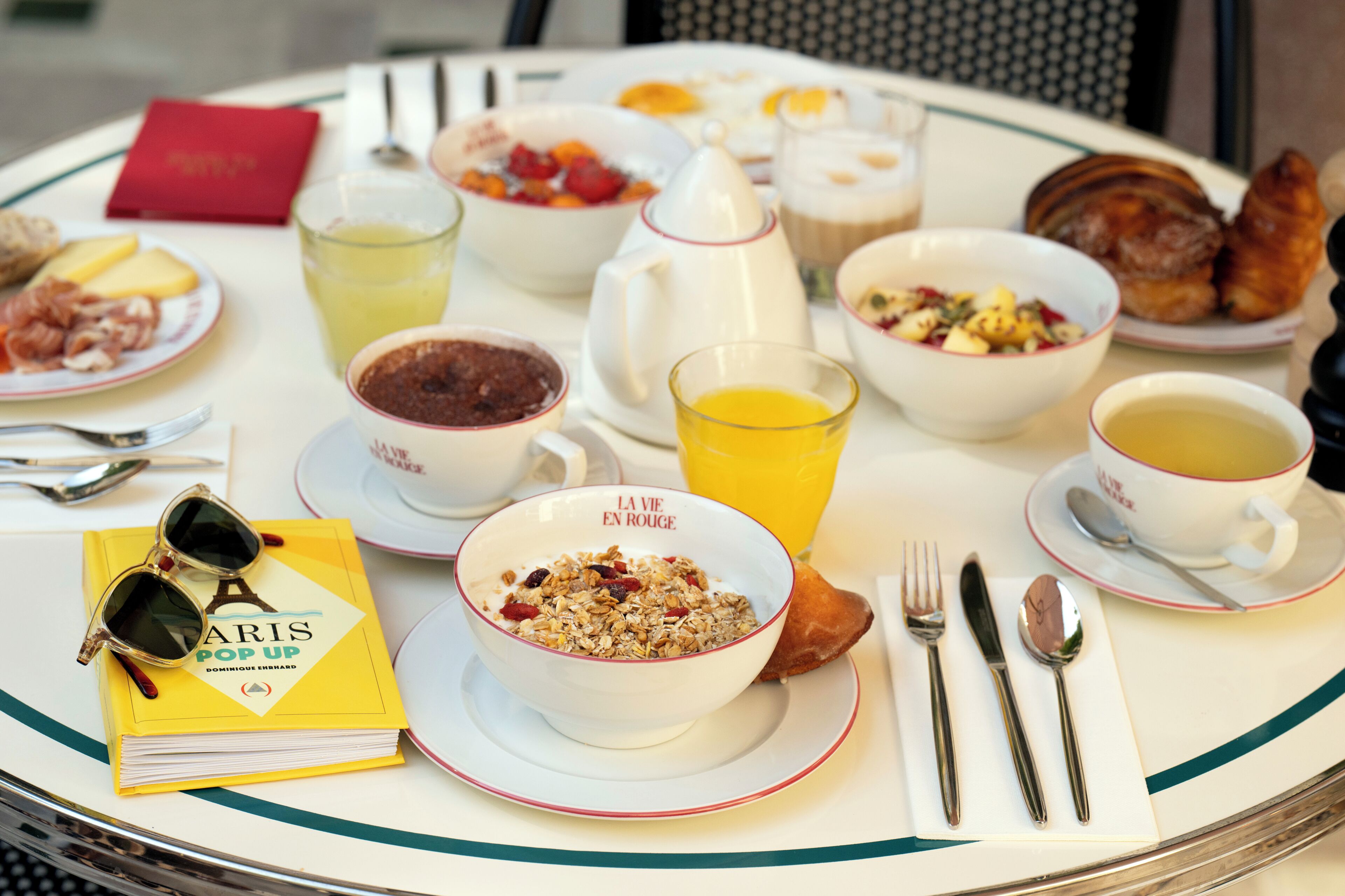 A breakfast setting with various dishes, a book, and a pair of sunglasses on a table.