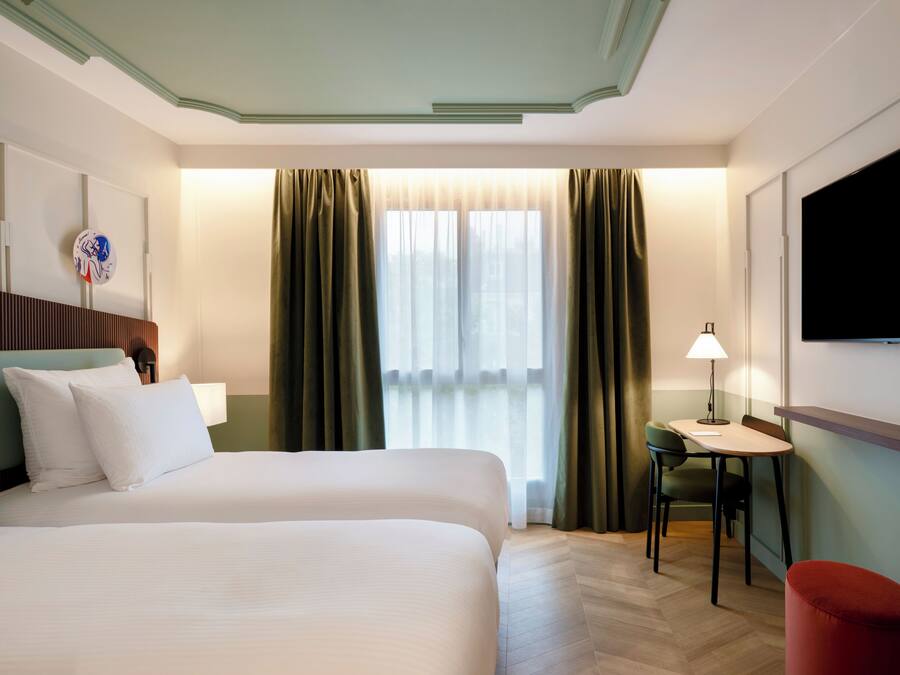 Habitación de hotel con cama, escritorio, silla, televisor y ventana con cortinas.