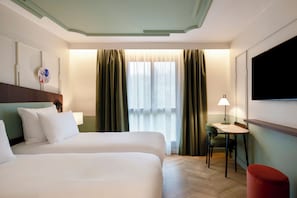 A hotel room with a bed, a desk, a chair, a TV, and a window with curtains. - Urban Hive Paris (Paris)
