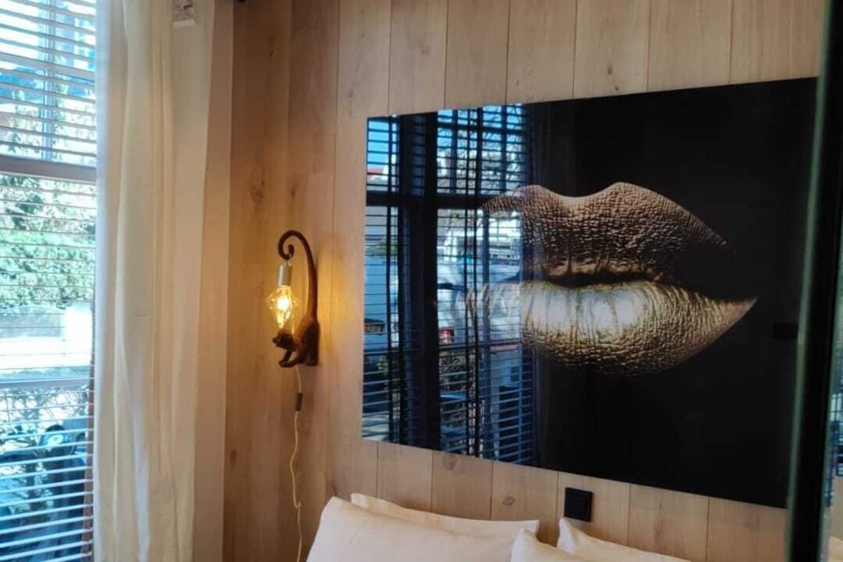 Ein Schlafzimmer mit Holzboden, einem Bett mit weißen Leinen, einer wandmontierten Leuchte und einem großen Spiegel, der den Raum reflektiert.