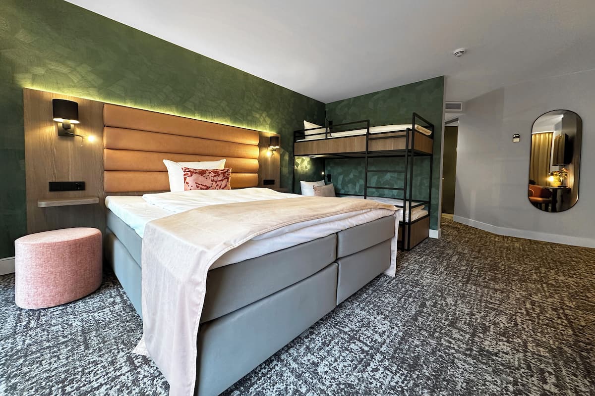 Una habitación de hotel moderna con una cama litera, una cama grande con colcha blanca, un otoman rosa y suelo alfombrado.