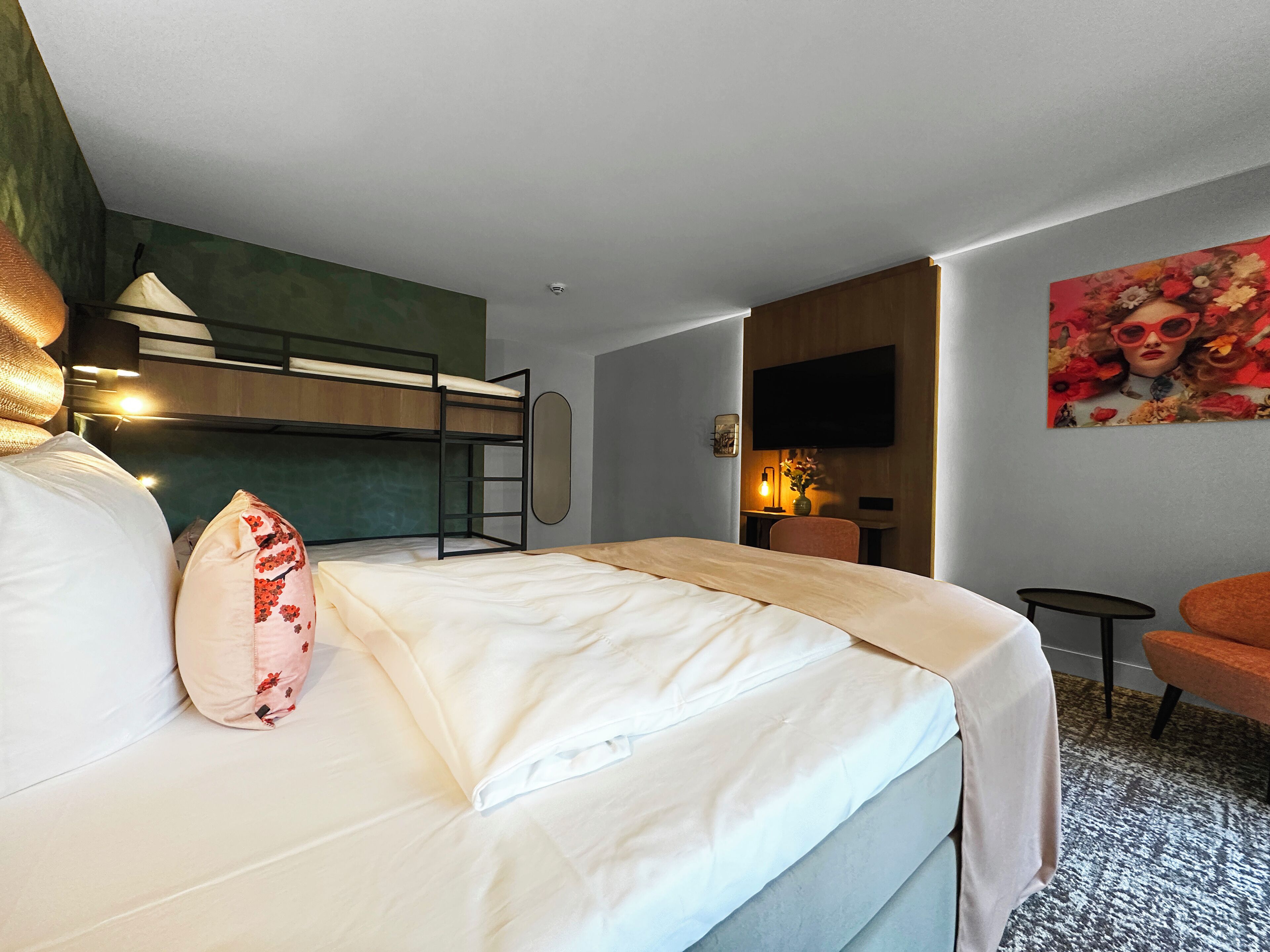 A hotel room with a bunk bed, a desk with a chair, a TV mounted on the wall, and a painting.