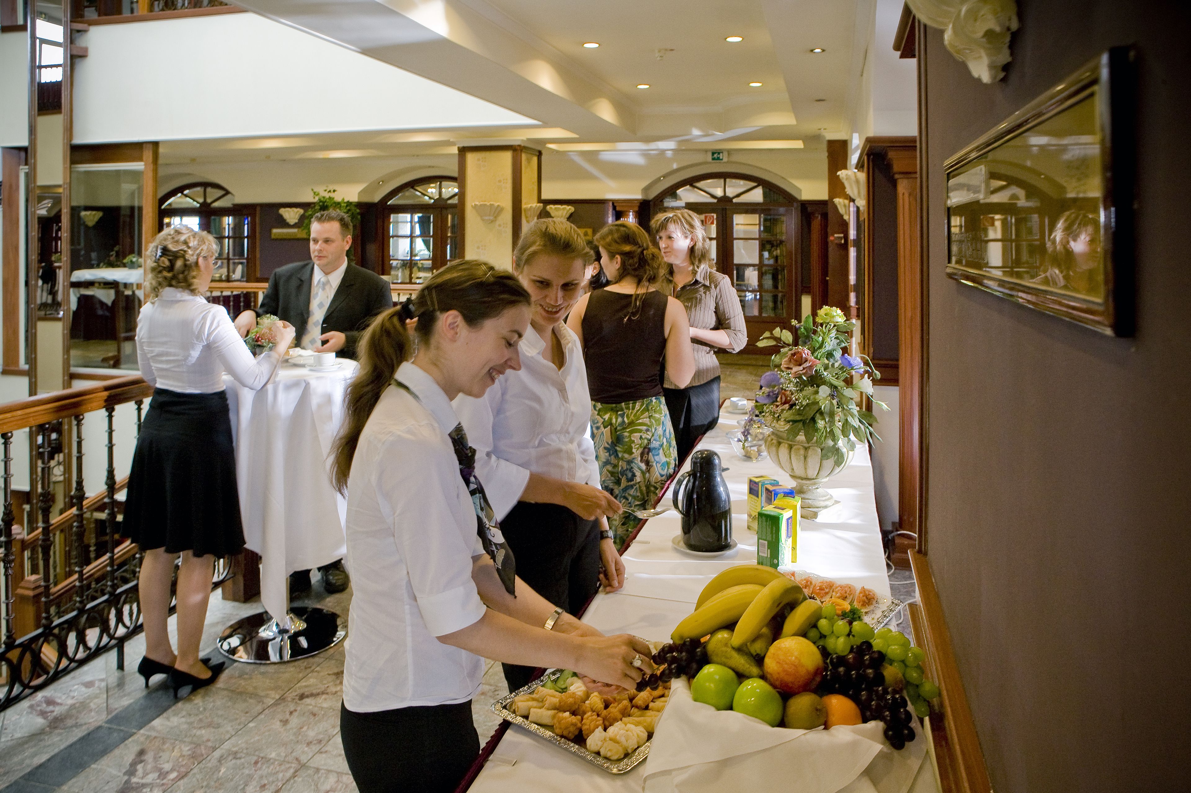 Un grupo de personas en un bufé con diversas frutas y una cafetera sobre la mesa.