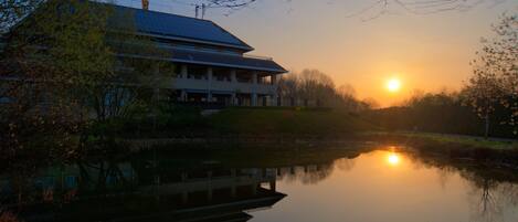 A building with a flat roof and a flagpole, situated next to a calm body of water reflecting the sunset.
