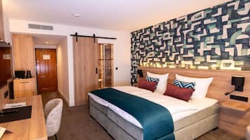 A hotel room with a bed, a desk, a chair, a wardrobe, and a wall with a geometric pattern.