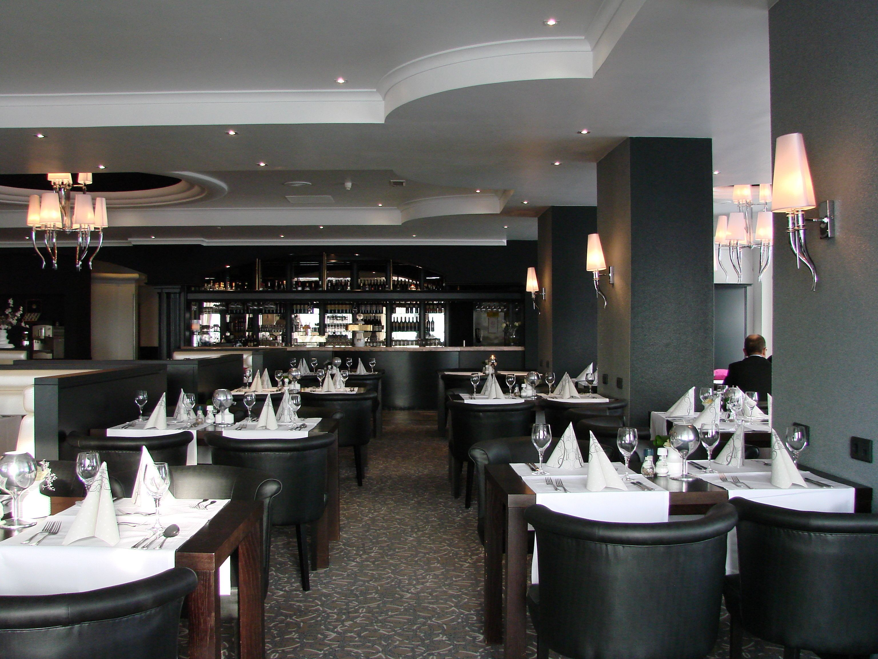 A restaurant with multiple tables set for dining, a bar area in the background, and a person seated at a table.