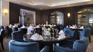 Un comedor con mesas redondas dispuestas para cuatro personas, sillas tapizadas en azul y un diseño interior moderno con paredes oscuras y ventanas amplias.