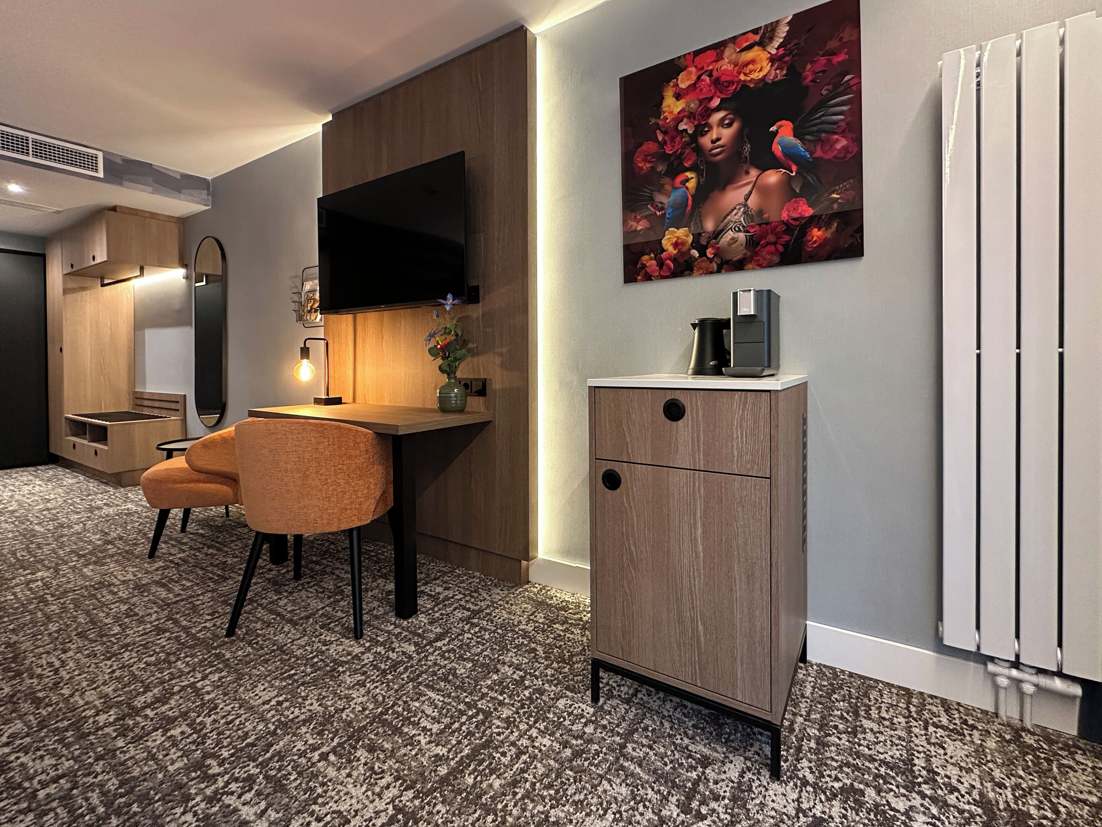 A modern hotel room with a wooden desk, a comfortable chair, a flat-screen TV mounted on the wall, a small cabinet with a coffee machine, and a radiator.