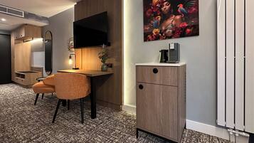 A modern hotel room with a wooden desk, a comfortable chair, a flat-screen TV mounted on the wall, a small cabinet with a coffee machine, and a radiator.
