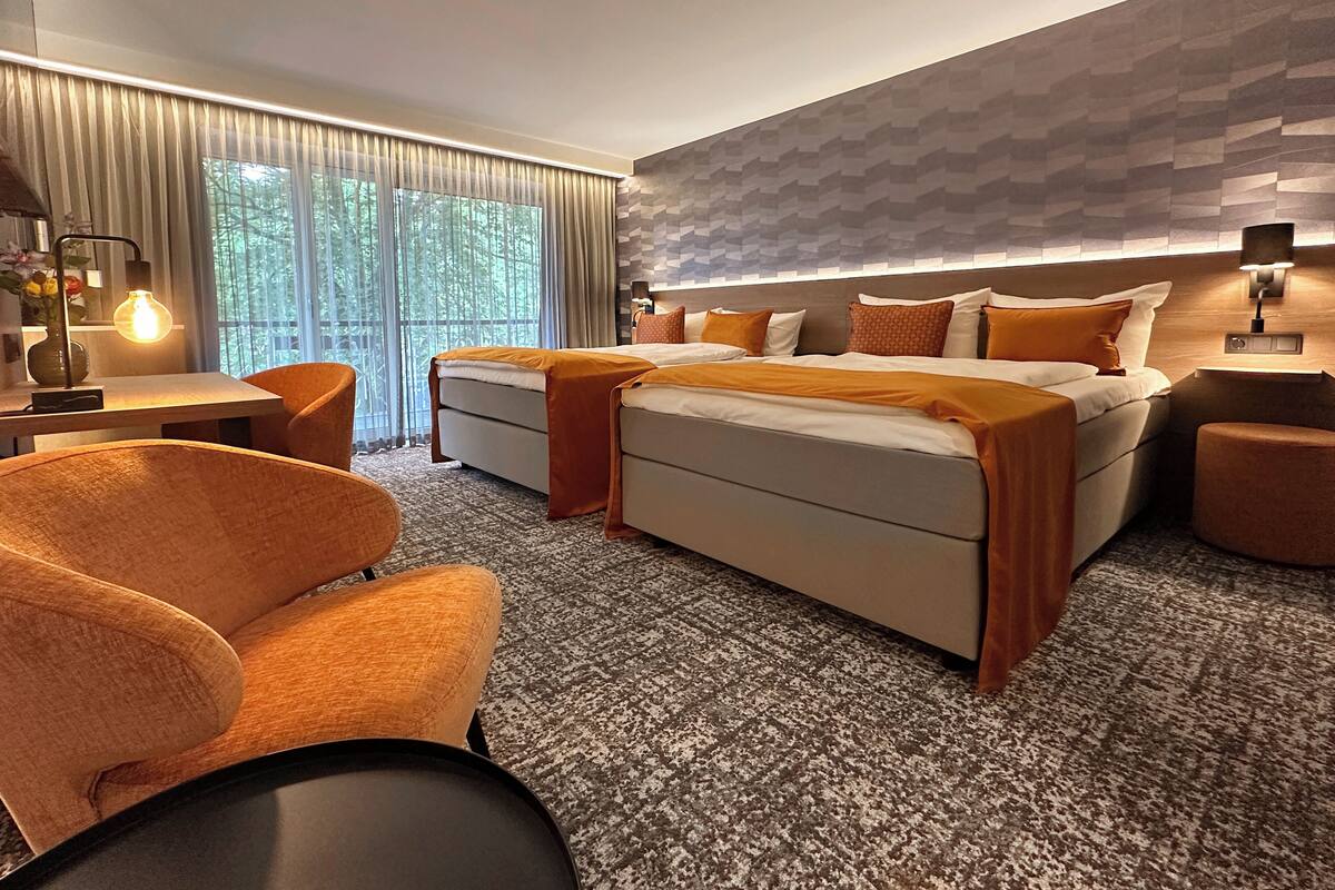 Habitación de hotel moderna con una cama grande, una zona de estar con una silla y una mesa, y vistas al exterior a través de amplios ventanales.