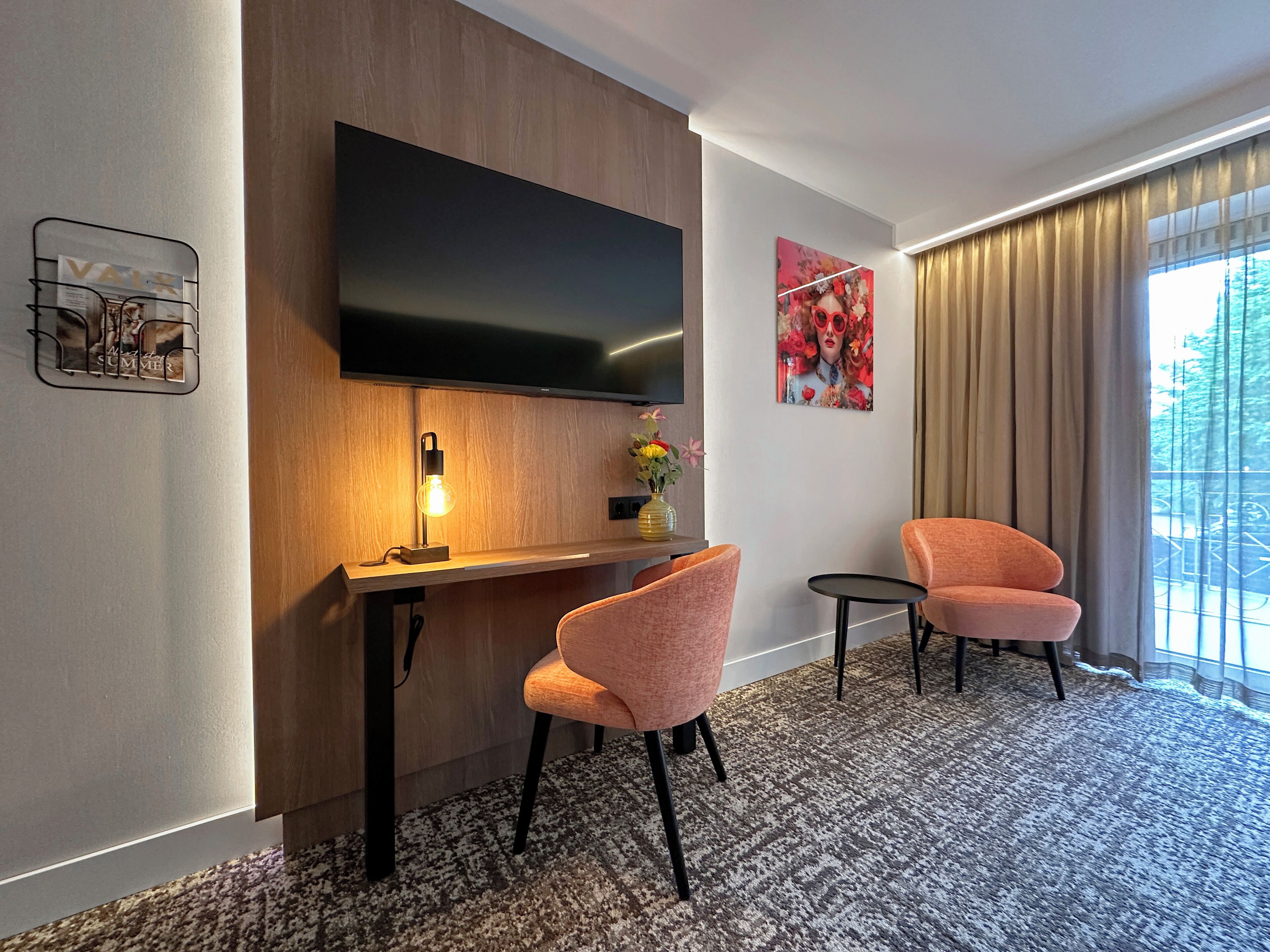 Una habitación de hotel moderna con una televisión de pantalla plana montada en la pared, un escritorio de madera con una lámpara, dos sillones rosados y una pequeña mesa redonda.
