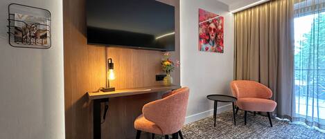 Una habitación de hotel moderna con una televisión de pantalla plana montada en la pared, un escritorio de madera con una lámpara, dos sillones rosados y una pequeña mesa redonda.