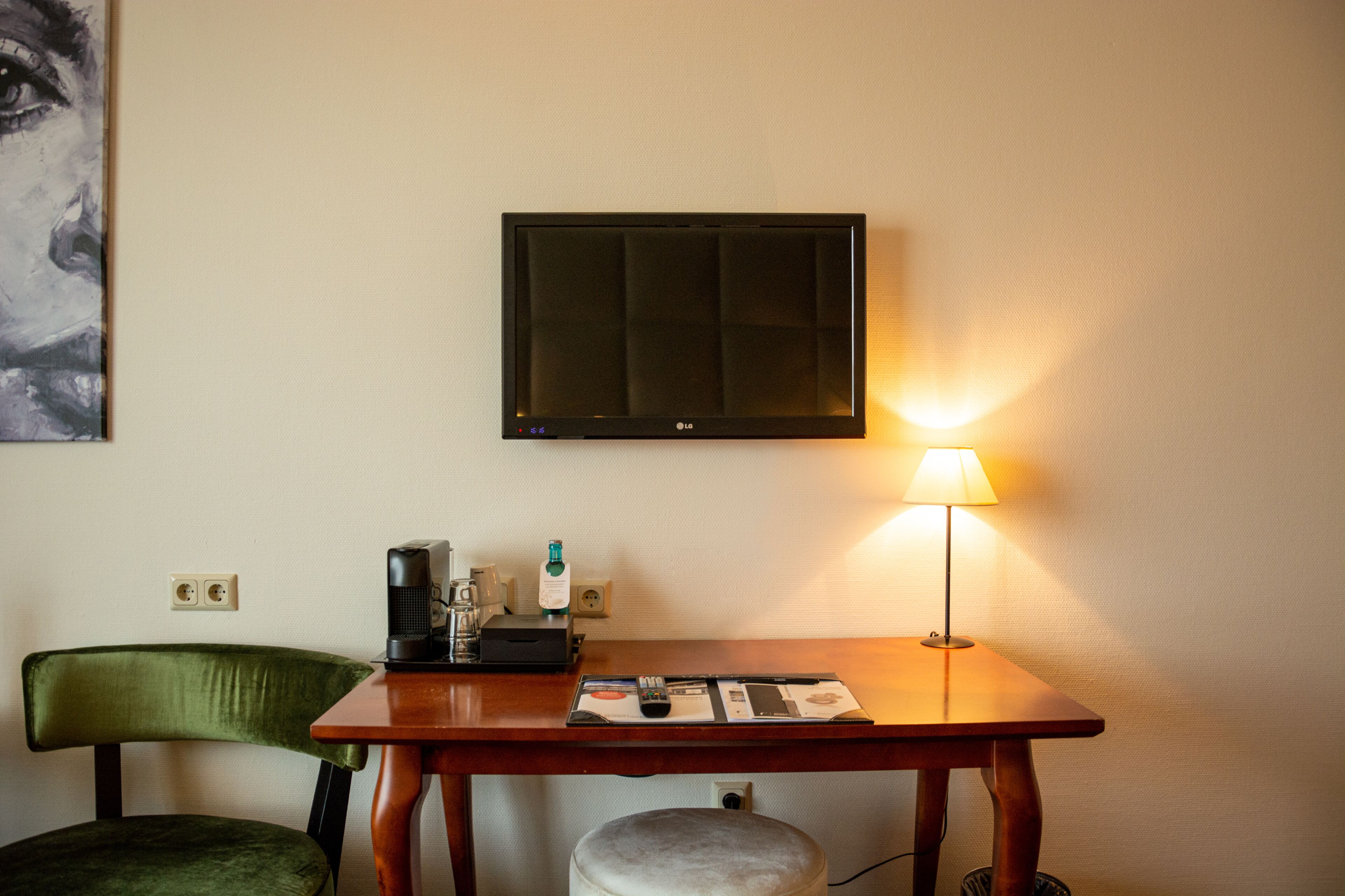 A hotel room with a flat-screen TV mounted on the wall, a table with a lamp, a chair, and a small table with a remote control and a magazine.