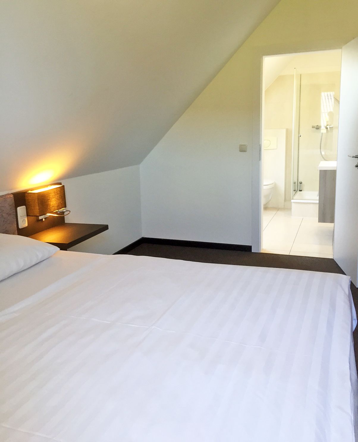 A bedroom with a bed, a nightstand with a lamp, and a view of a bathroom through an open door.