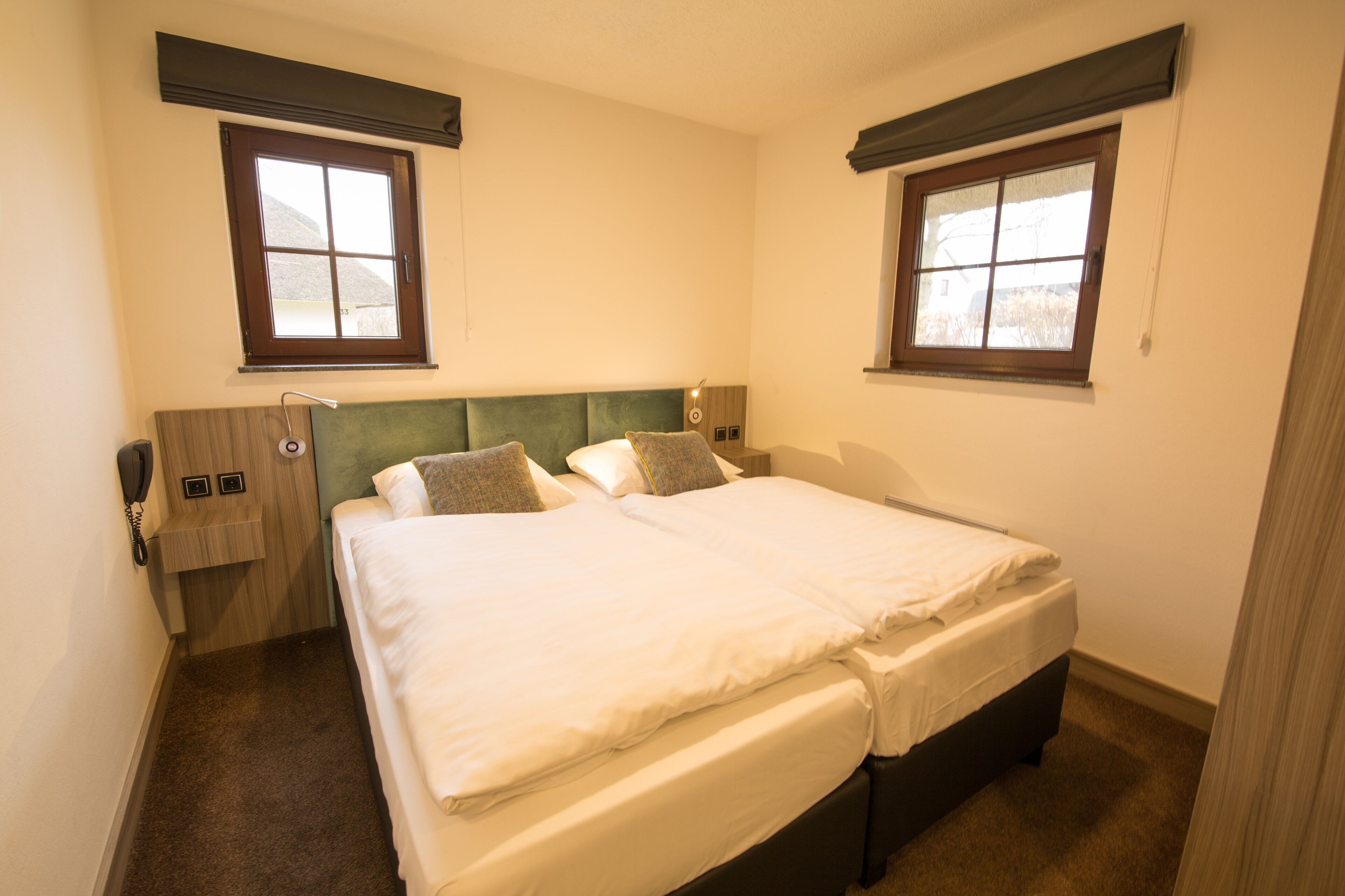 Une chambre d’hôtel avec un lit double, deux tables de chevet, un téléphone et deux fenêtres équipées de stores.