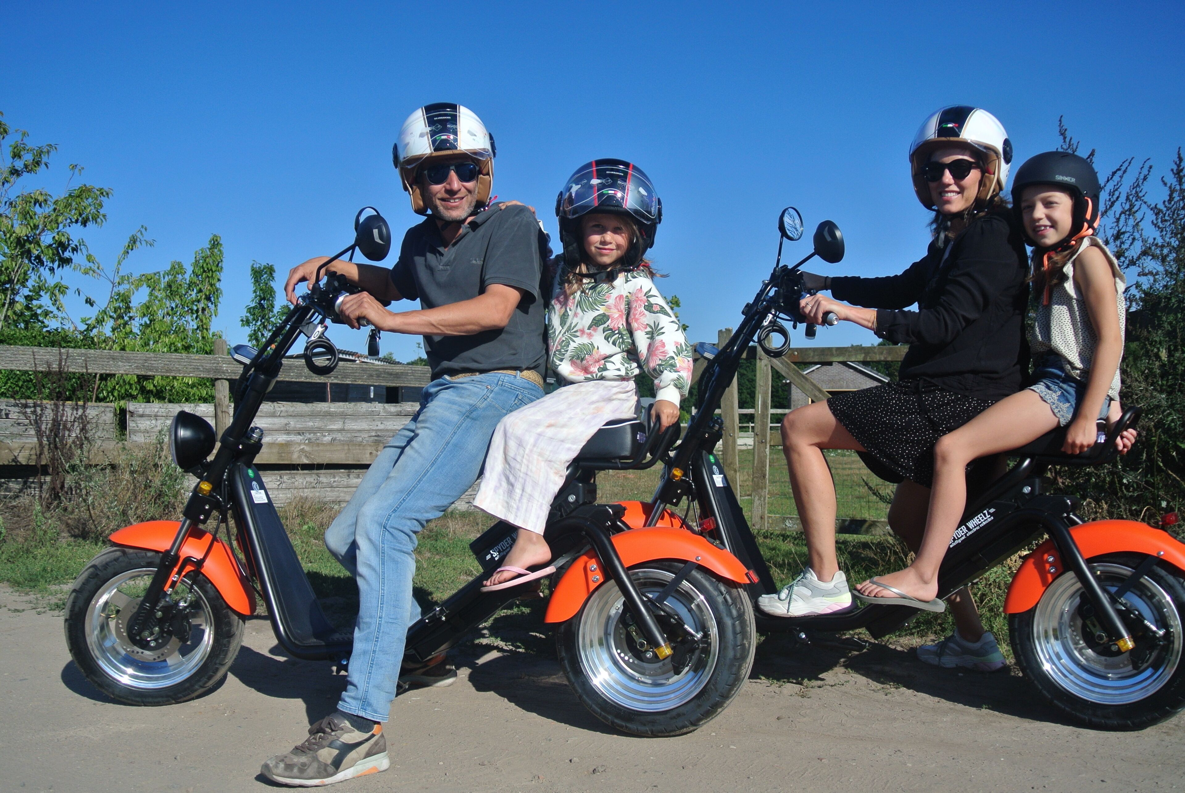 Une famille fait du vélo sur un tandem, avec un ciel dégagé et une végétation luxuriante en arrière-plan.