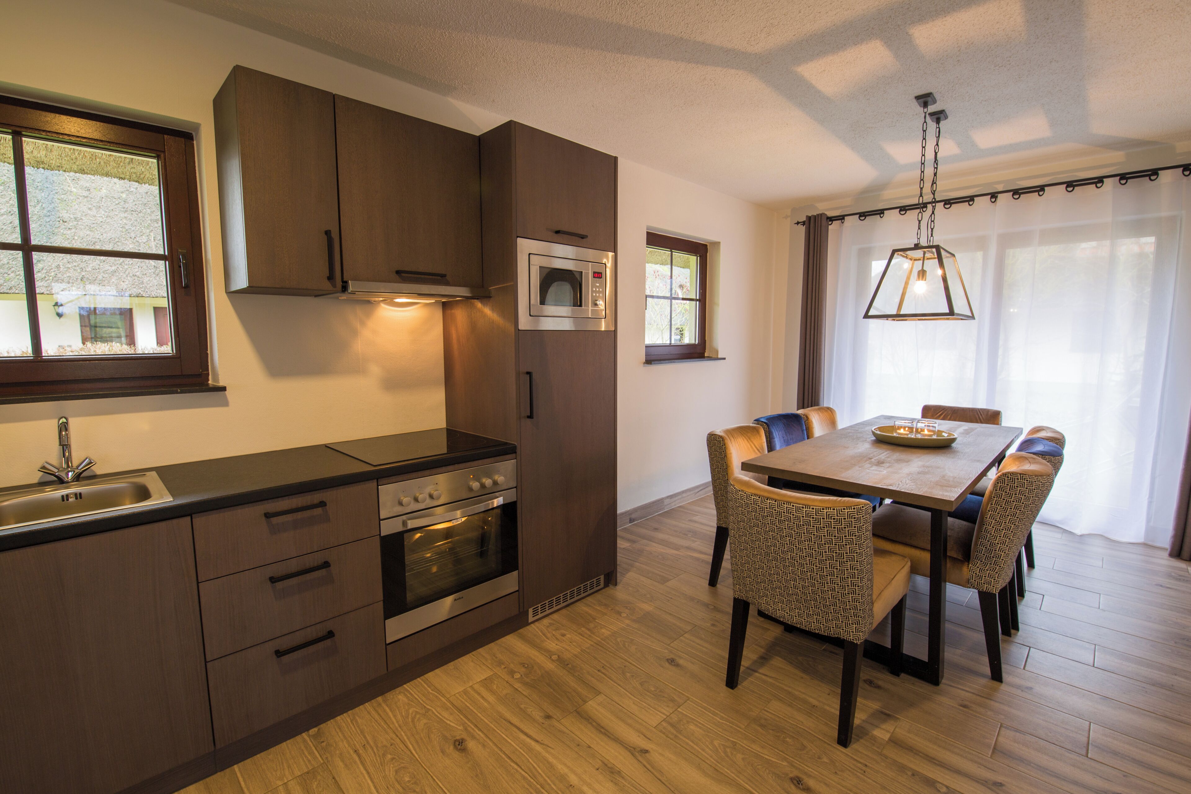 A modern kitchen with built-in appliances, a dining area with a wooden table and chairs, and large windows with curtains.