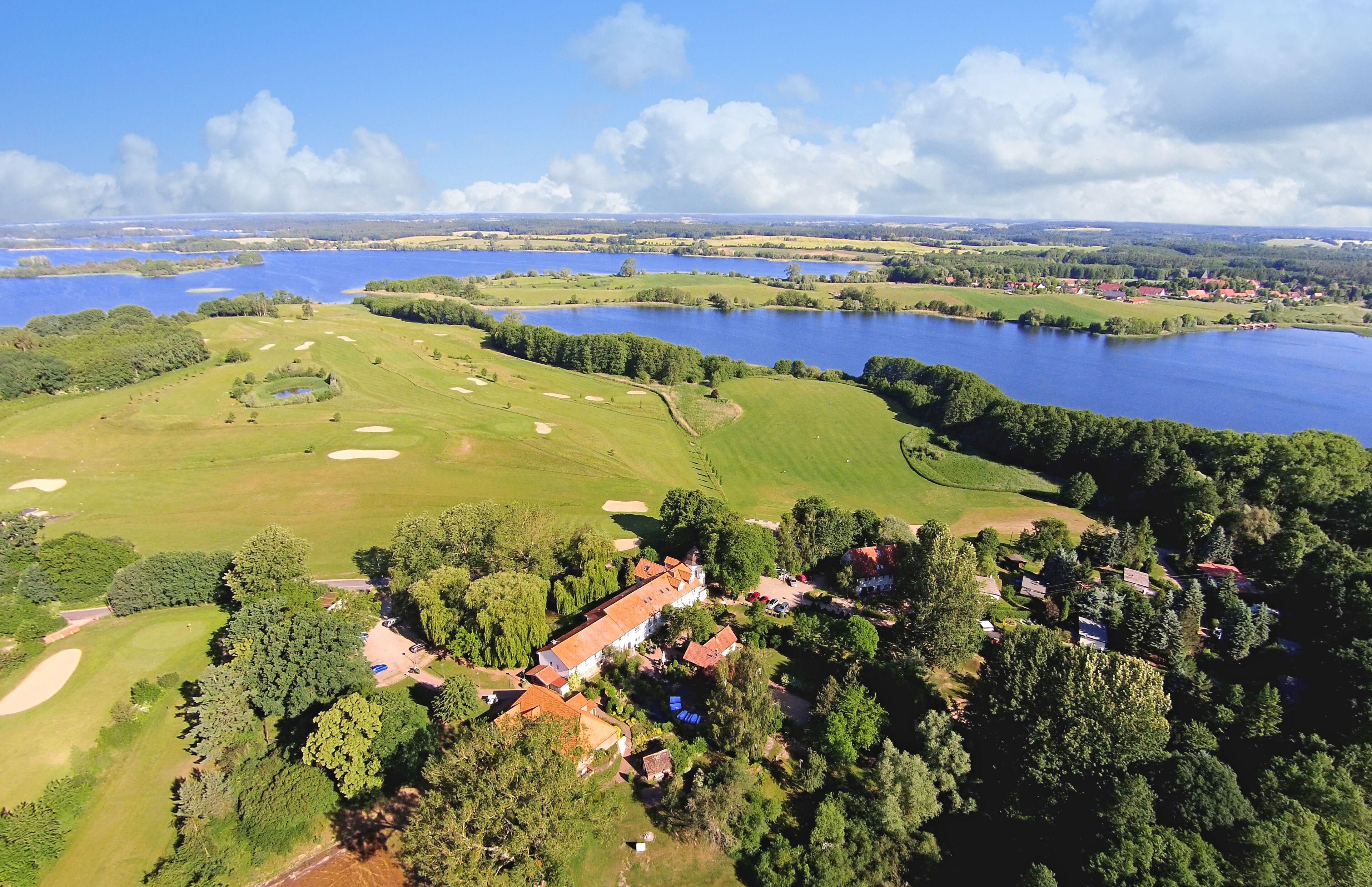 An aerial view of a golf course with a clubhouse, surrounded by water bodies and greenery.