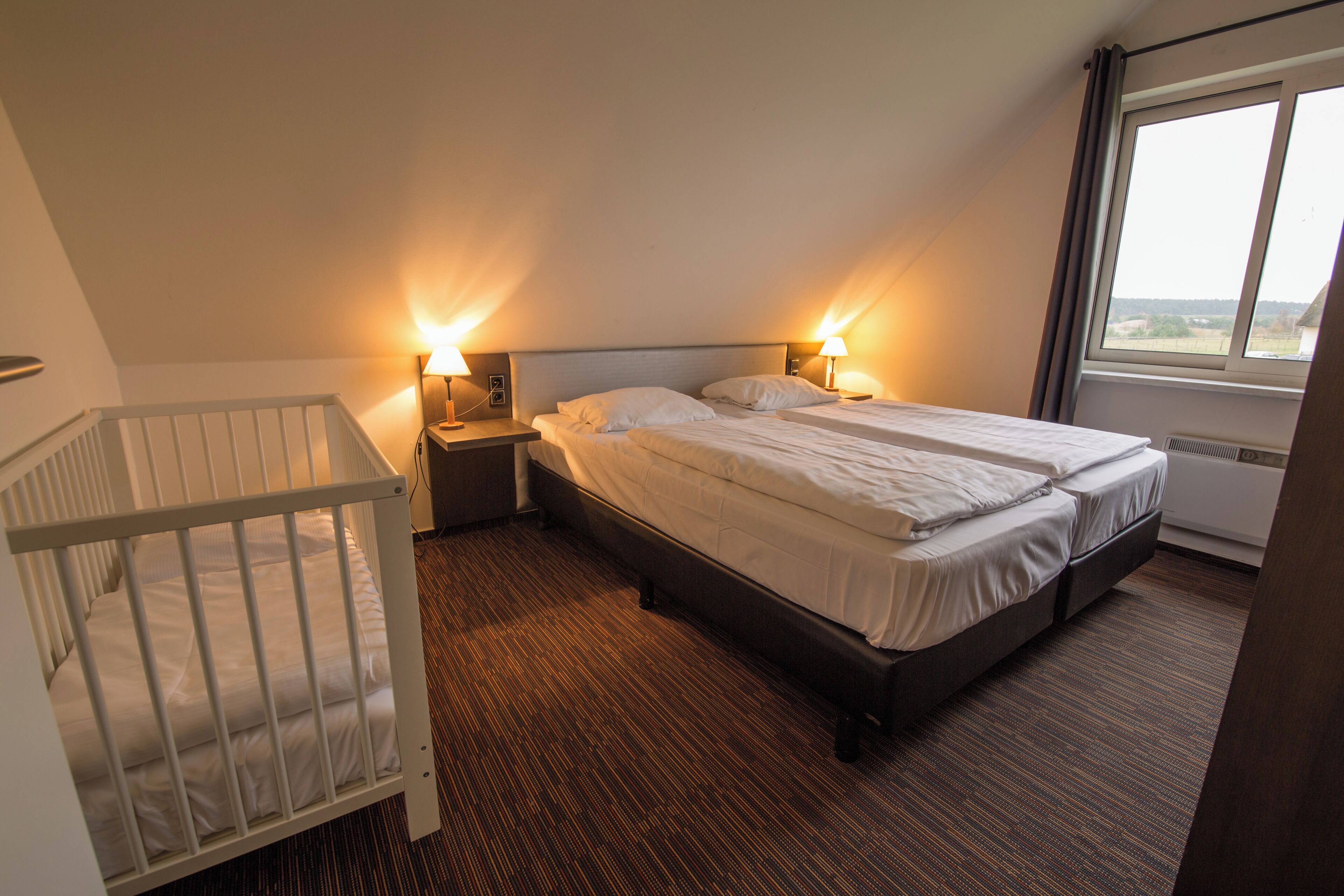 A hotel room with a crib, a double bed, two bedside lamps, and a window with a view.