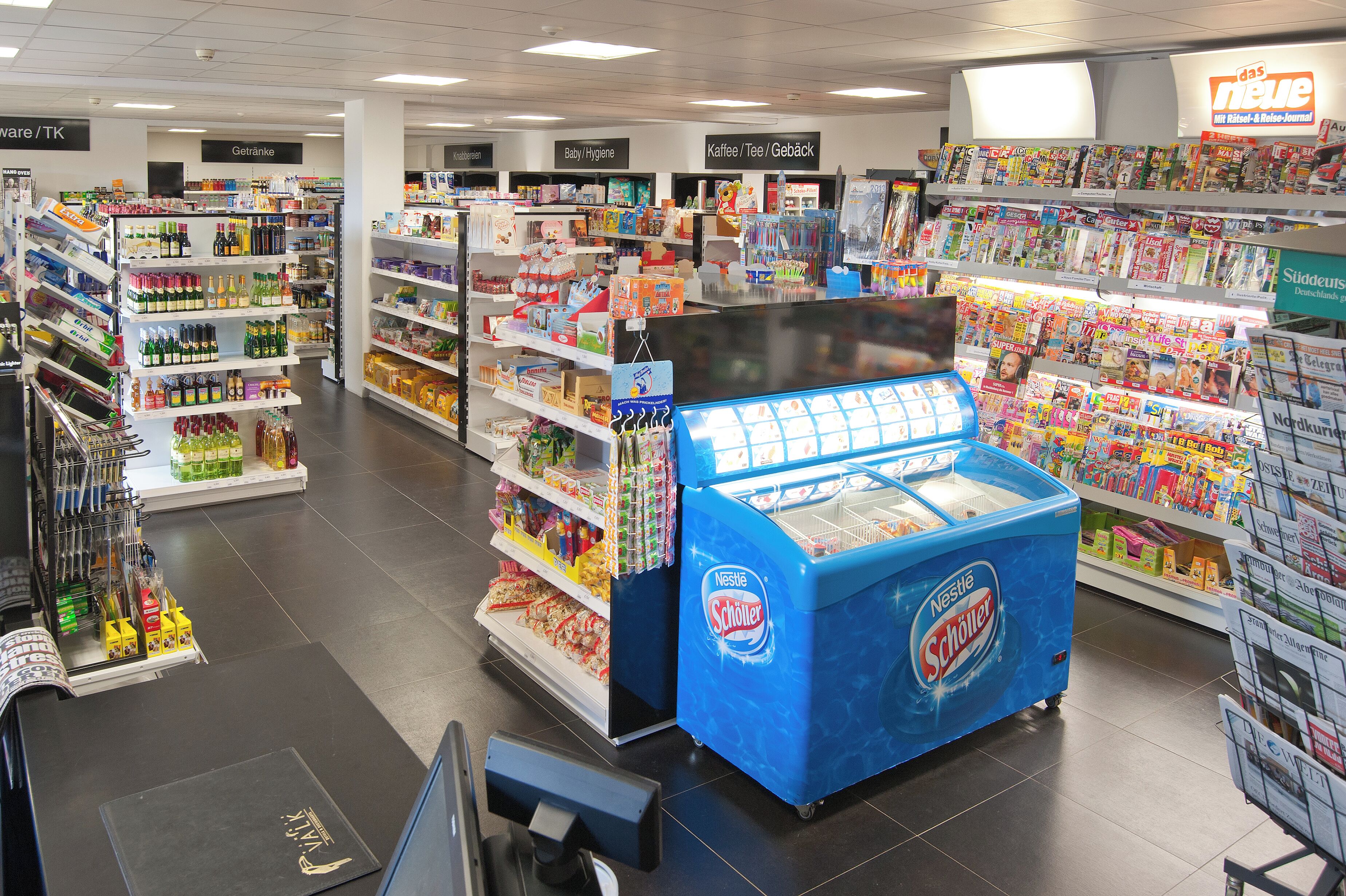 Un pasillo de supermercado con estantes repletos de diversos productos, un congelador de helados Nestlé Schöner y una zona de cajas.