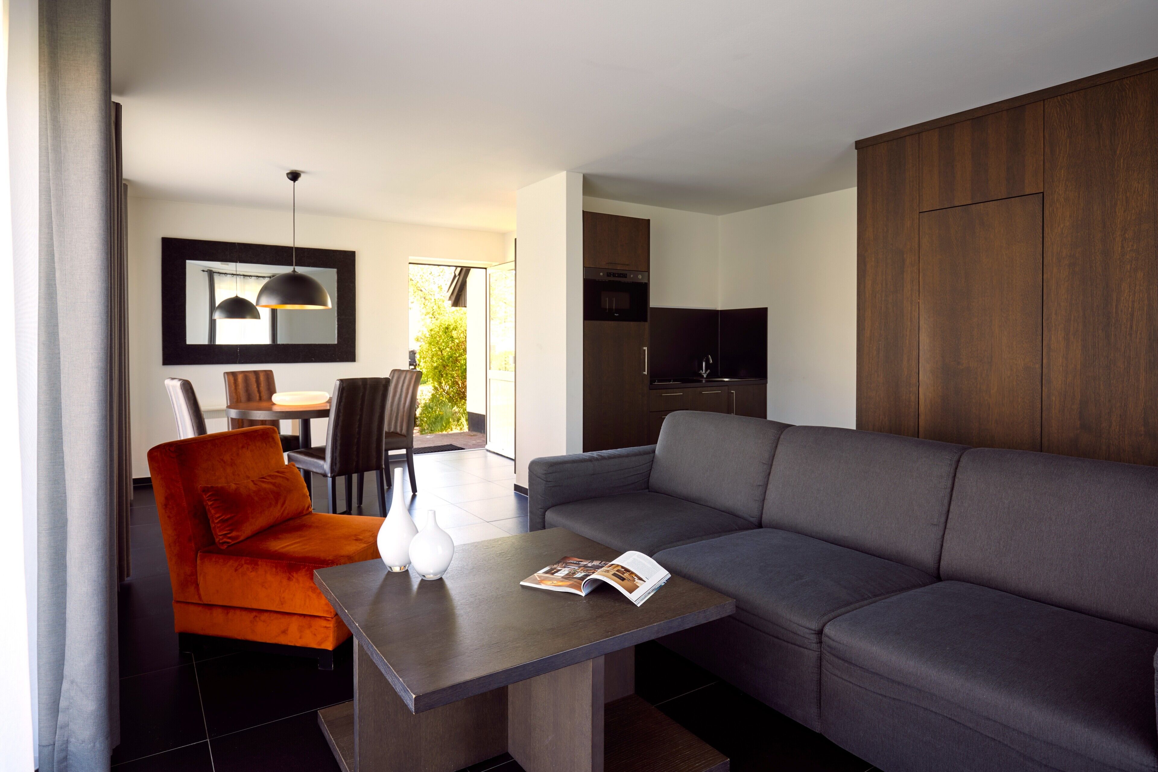 A modern living room with a grey sofa, orange armchair, and a dining area with a table and chairs.