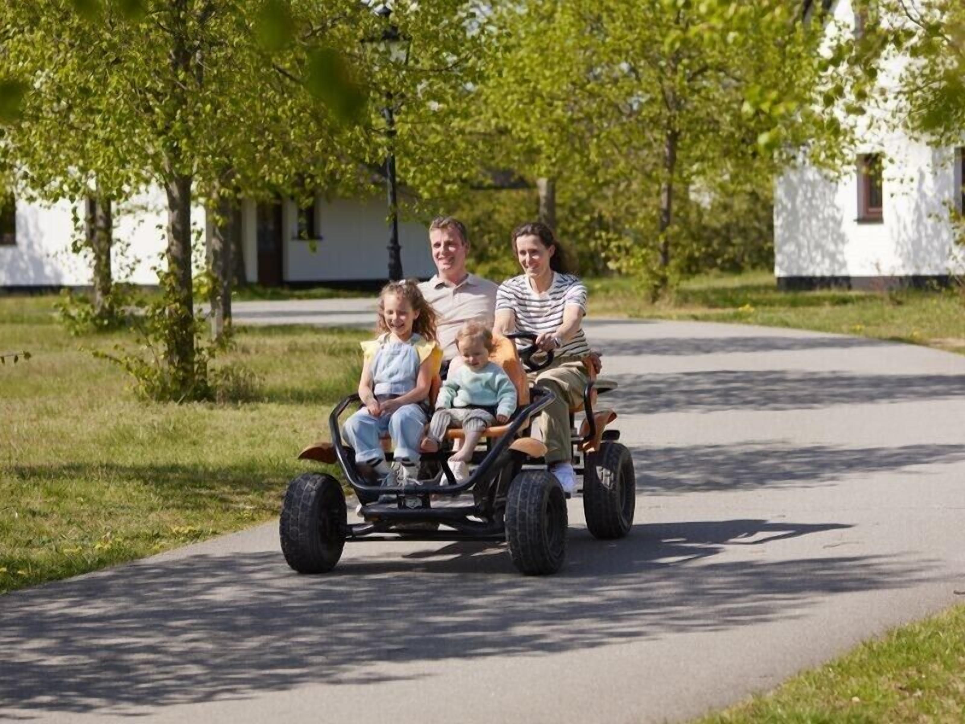 A family in a three-wheeled vehicle on a paved path with trees and white buildings in the background.