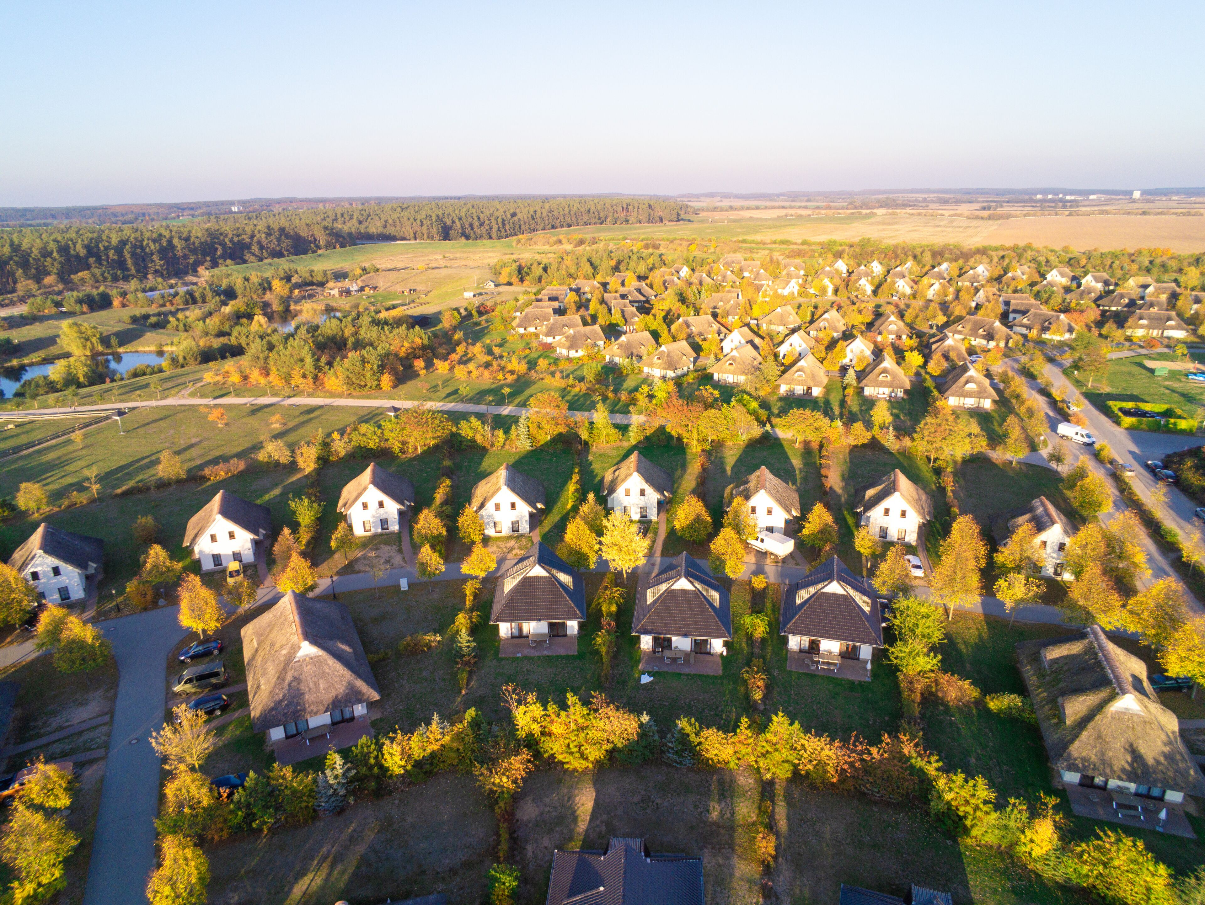 Vue aérienne d’un quartier résidentiel avec des maisons, dont certaines sont coiffées de toits de chaume, entourées d’arbres et de champs ouverts.