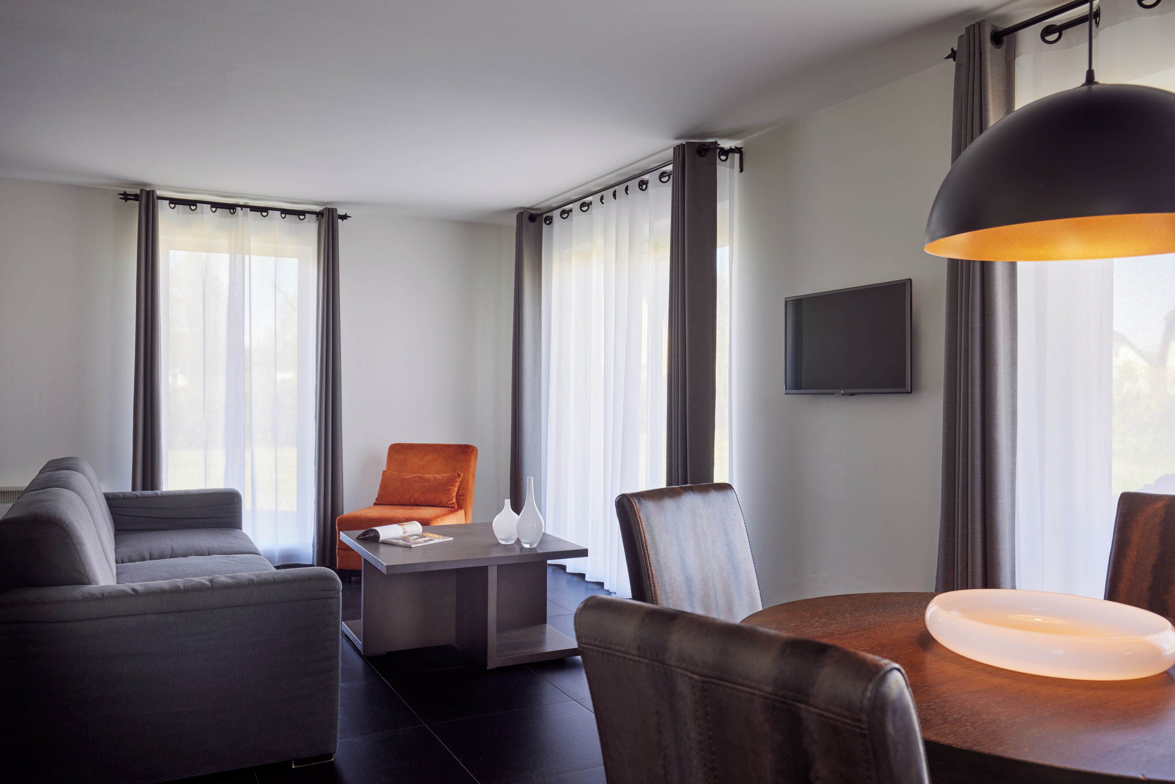 A modern hotel room with a sofa, a dining table, a chair, and a TV mounted on the wall.