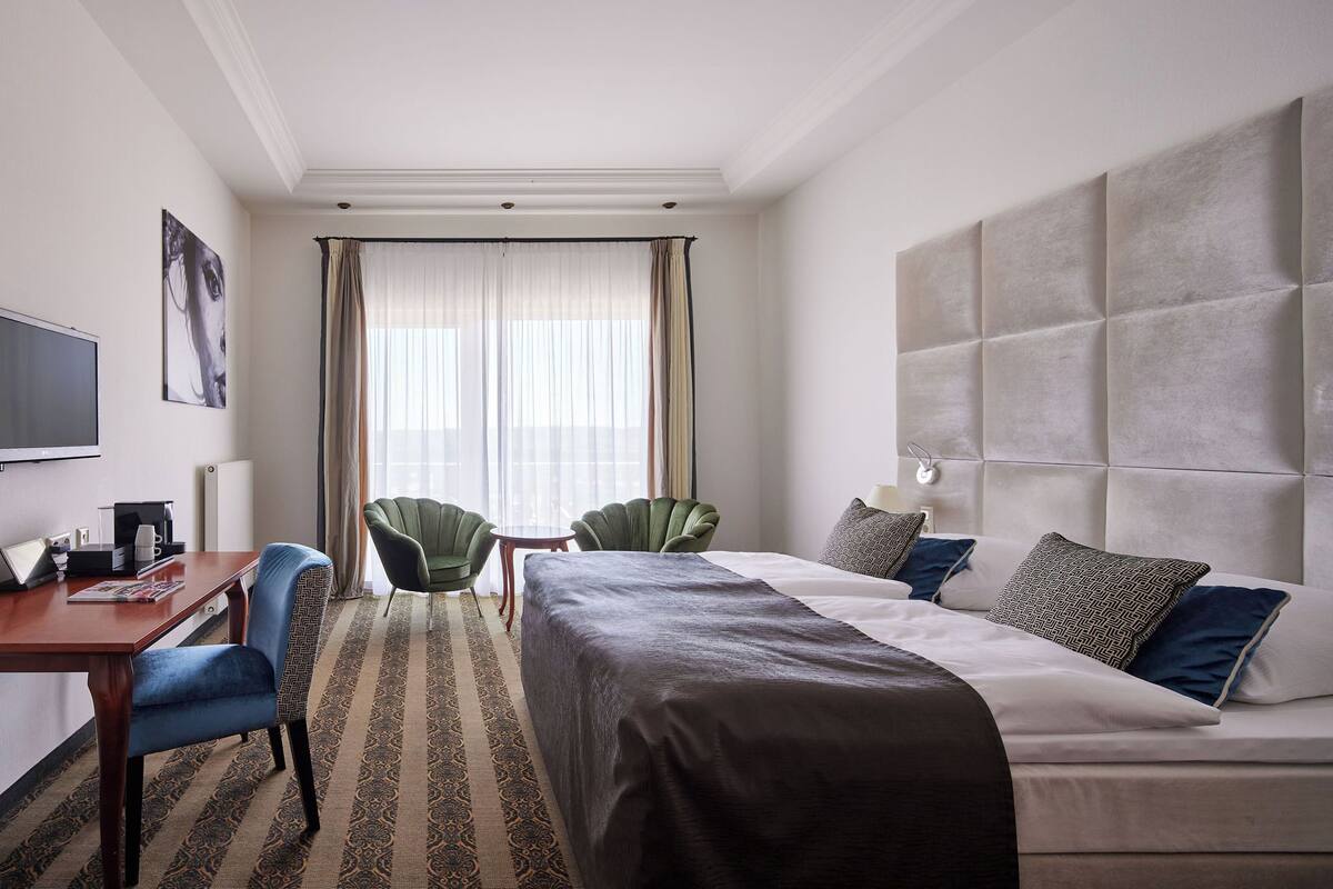 A hotel room with a bed, a desk, two chairs, a television, and a large window with curtains.