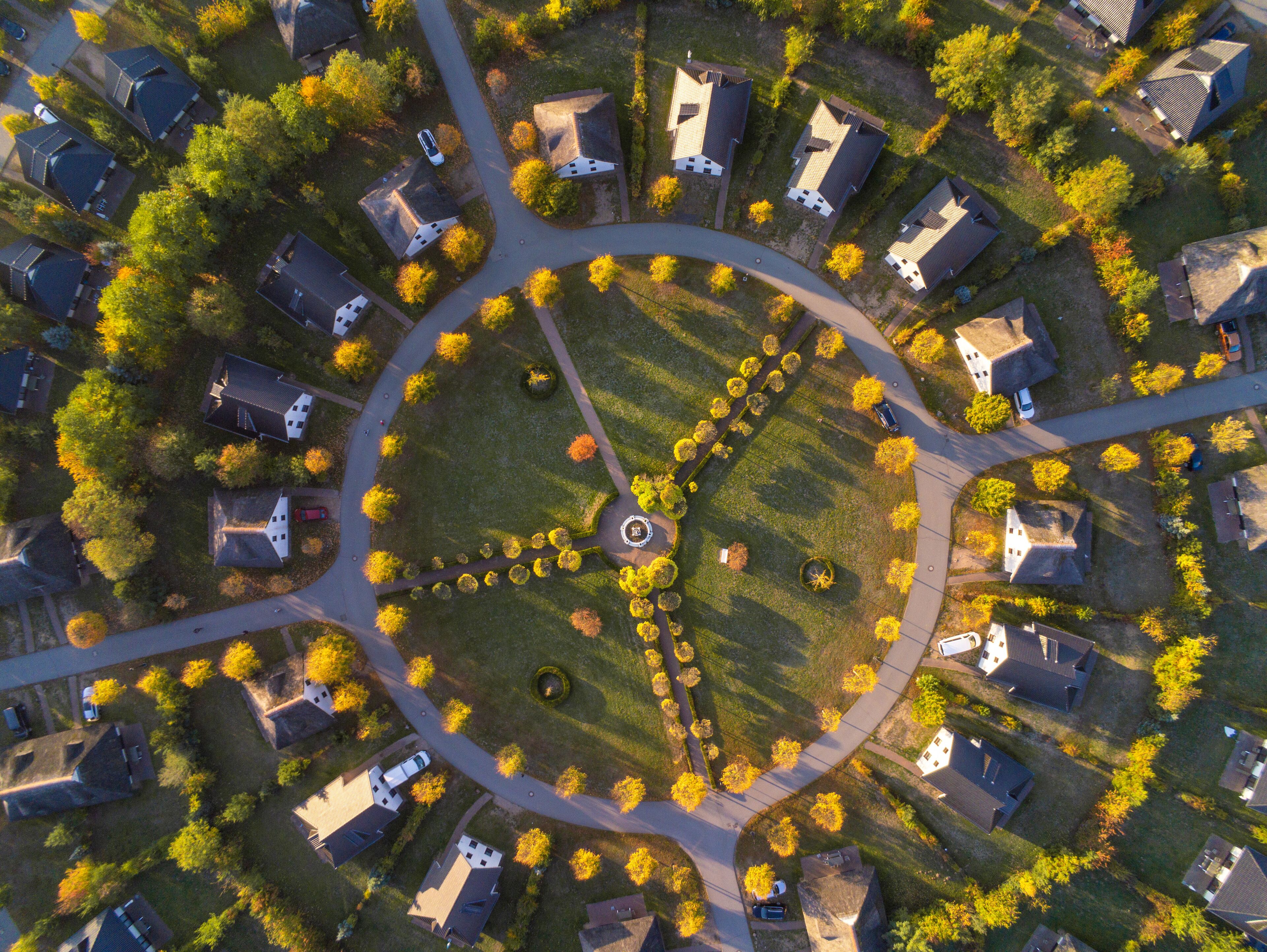 An aerial view of a residential area with houses arranged around a central circular park, featuring a fountain and a circular walking path.