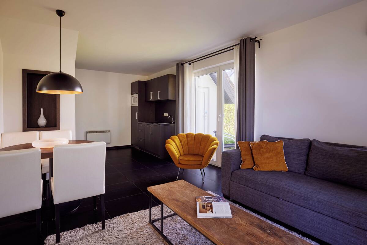 A modern living room with a sofa, a coffee table, a dining table with chairs, a pendant light, and a kitchen area in the background.