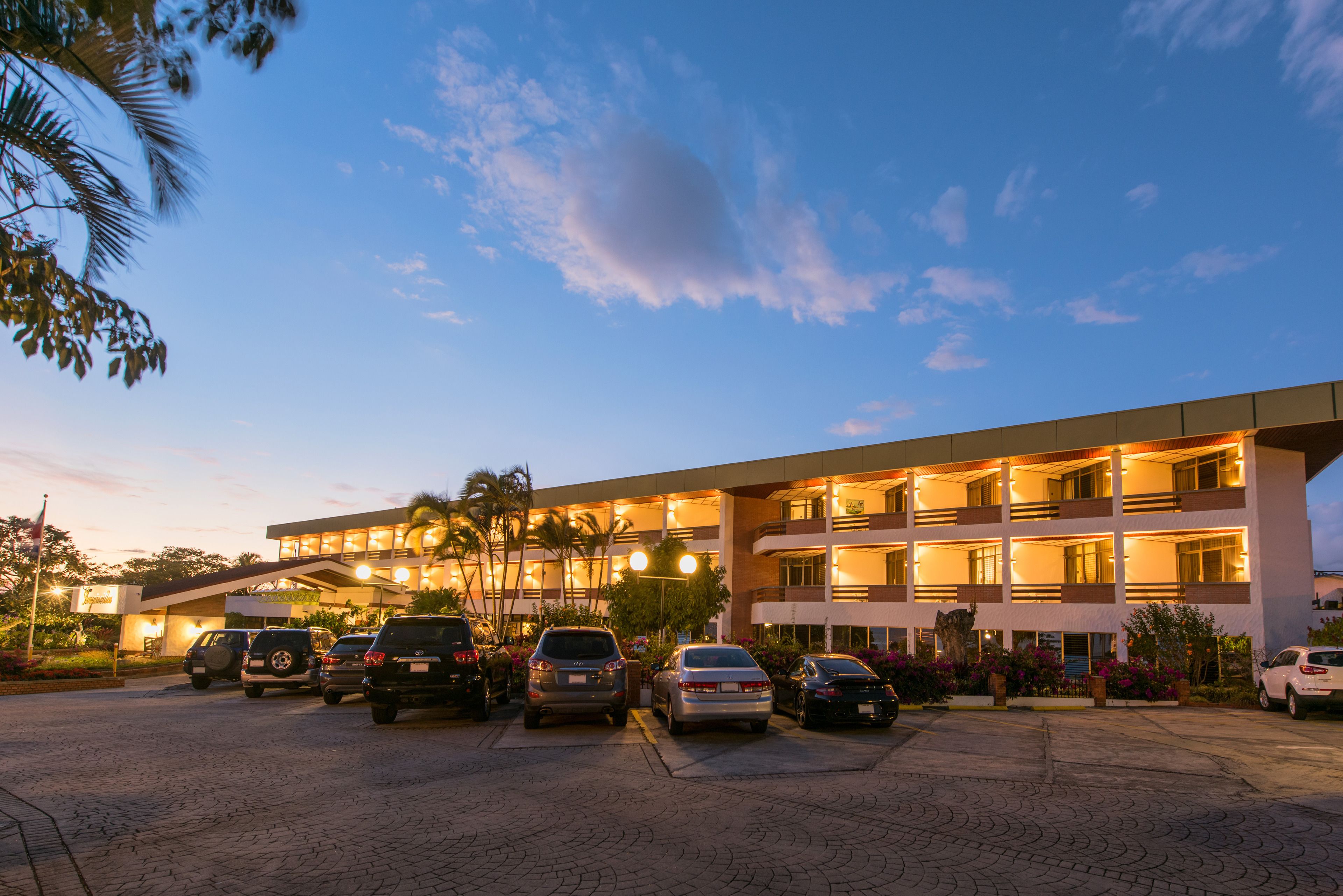 Un hotel de varios pisos con un estacionamiento lleno de coches, rodeado de palmeras e iluminado con una luz ambiental.