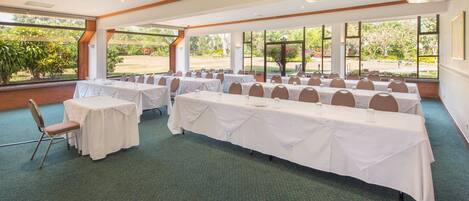 A conference room with multiple tables and chairs, large windows, and a view of greenery outside.