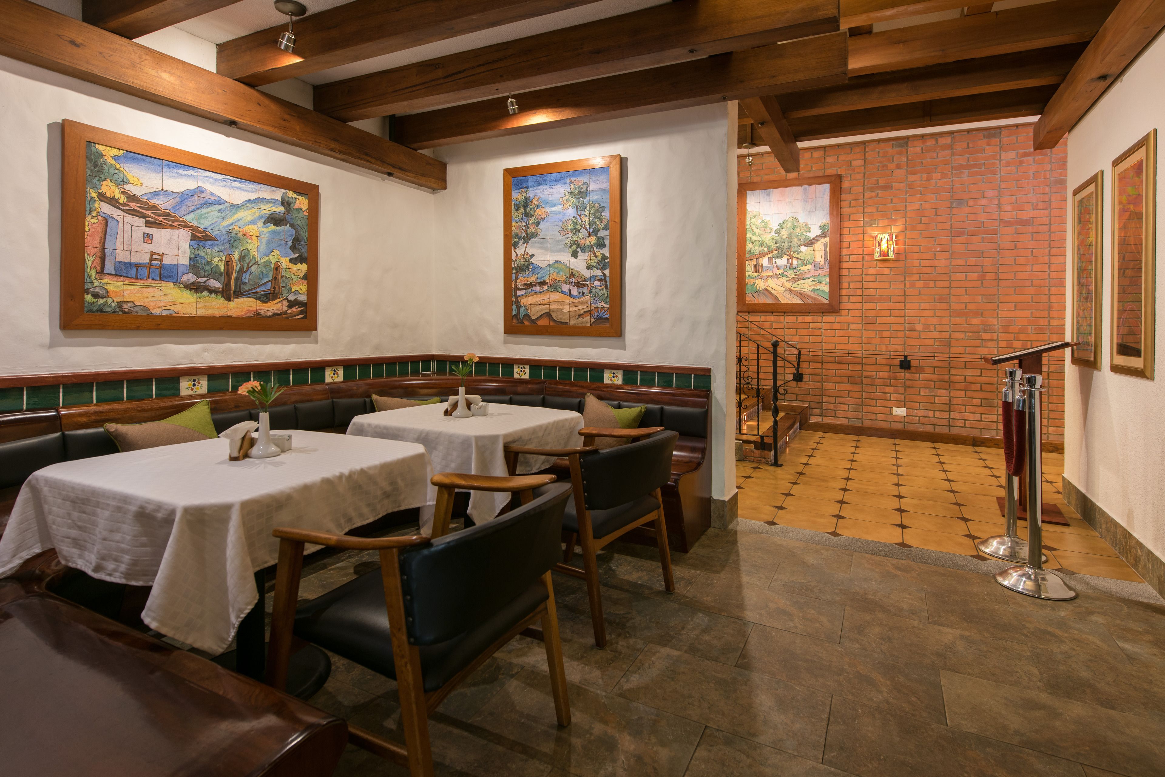 Restaurante com vigas de madeira, pisos ladrilhados e obras de arte nas paredes. Mesas montadas com toalhas brancas e cadeiras.