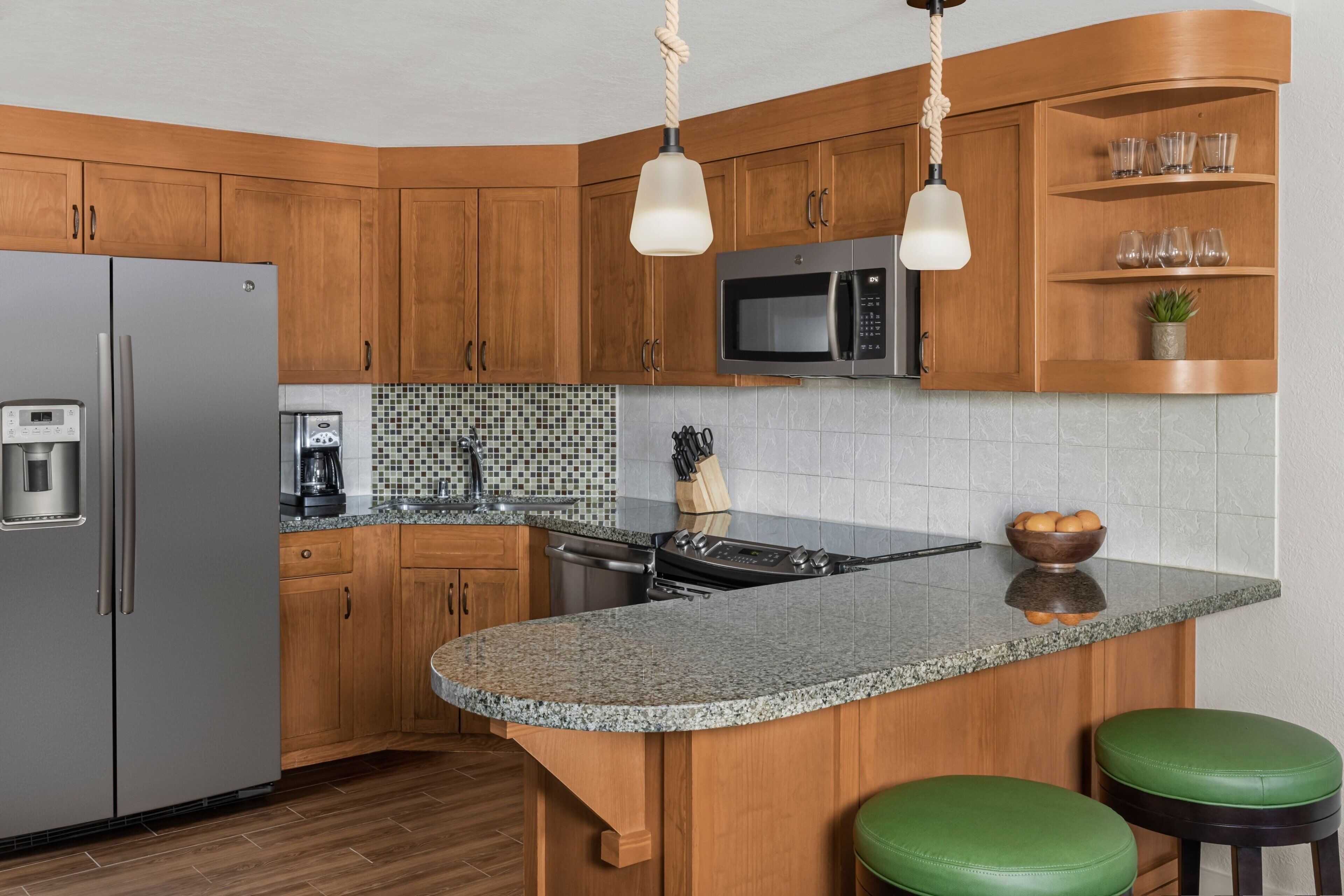 A kitchen with wooden cabinets, a stainless steel refrigerator, a microwave, a granite countertop, and two green stools.