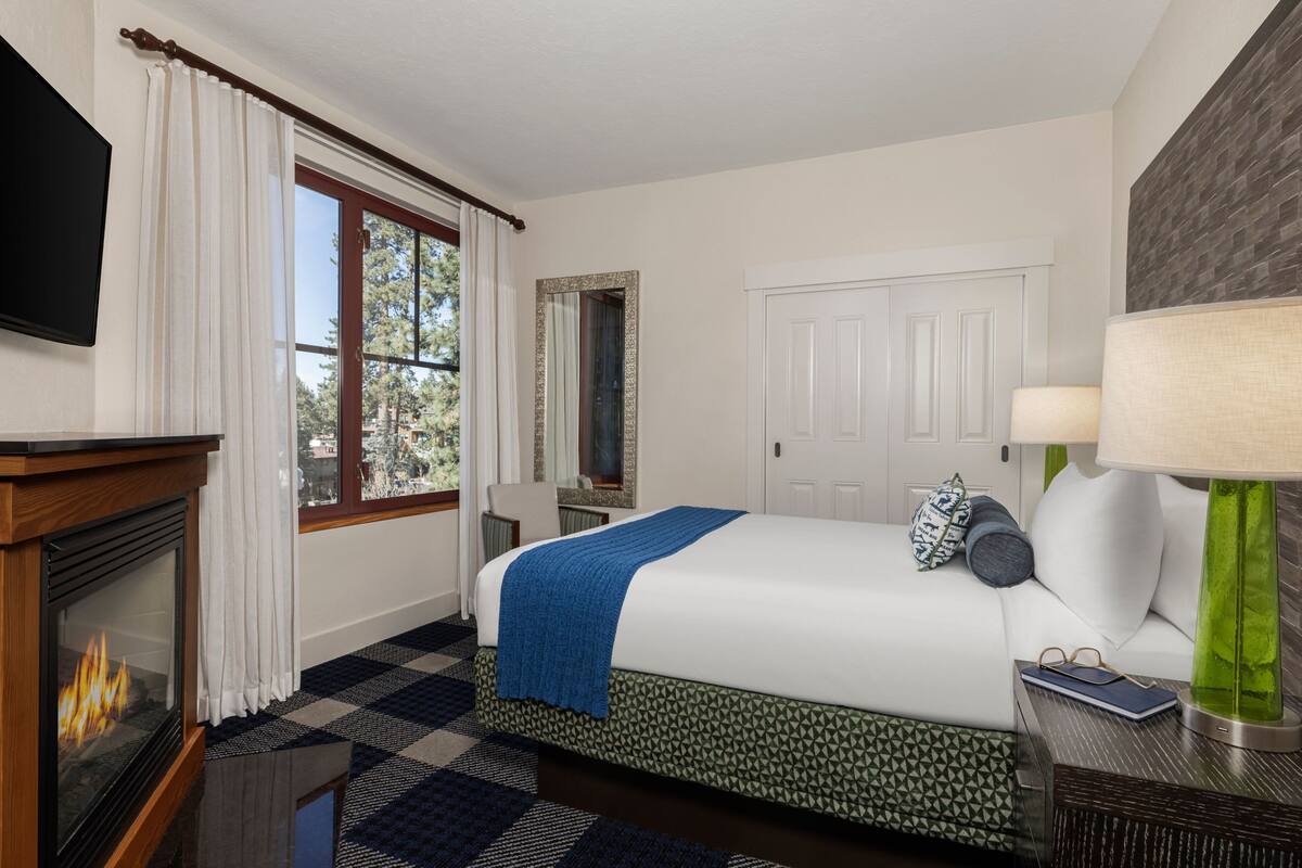 A hotel room with a bed, a fireplace, a window with curtains, a mirror, a door, and a lamp.