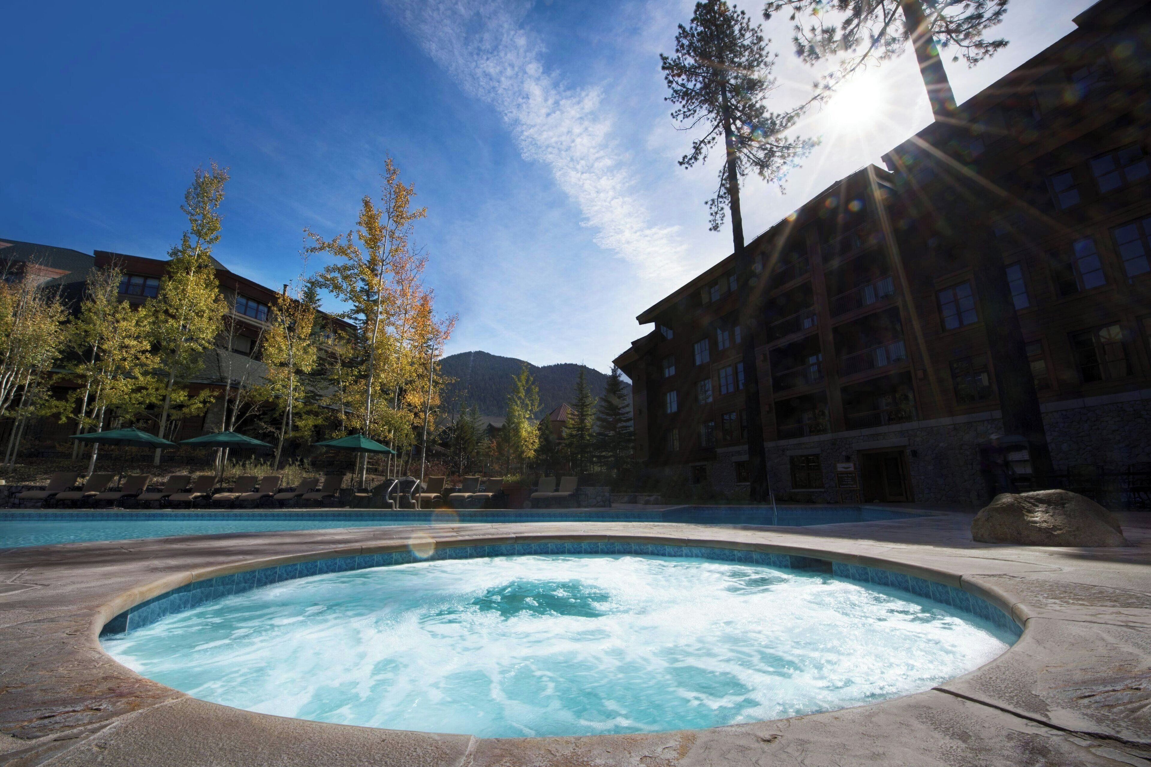 A hot tub surrounded by lounge chairs and umbrellas, with a pool in the background and a mountain in the distance.