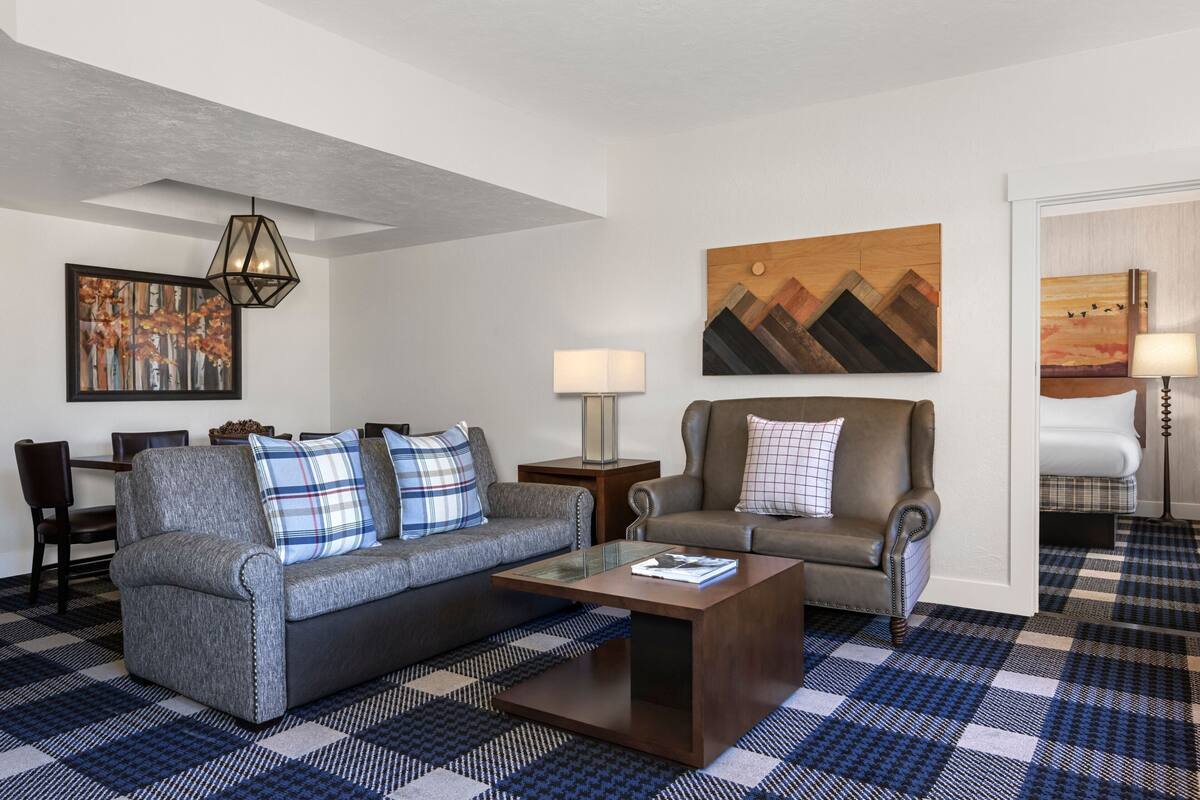 A modern living room with a gray sofa, a brown armchair, and a coffee table. There are two side tables with lamps, a painting on the wall, and a checkered floor.