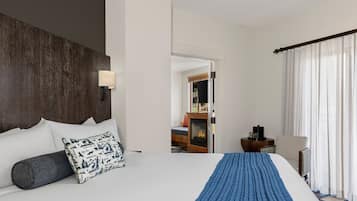 Quarto de hotel com uma cama grande, uma mesa de cabeceira, uma cadeira e vista para uma lareira através de uma porta aberta.
