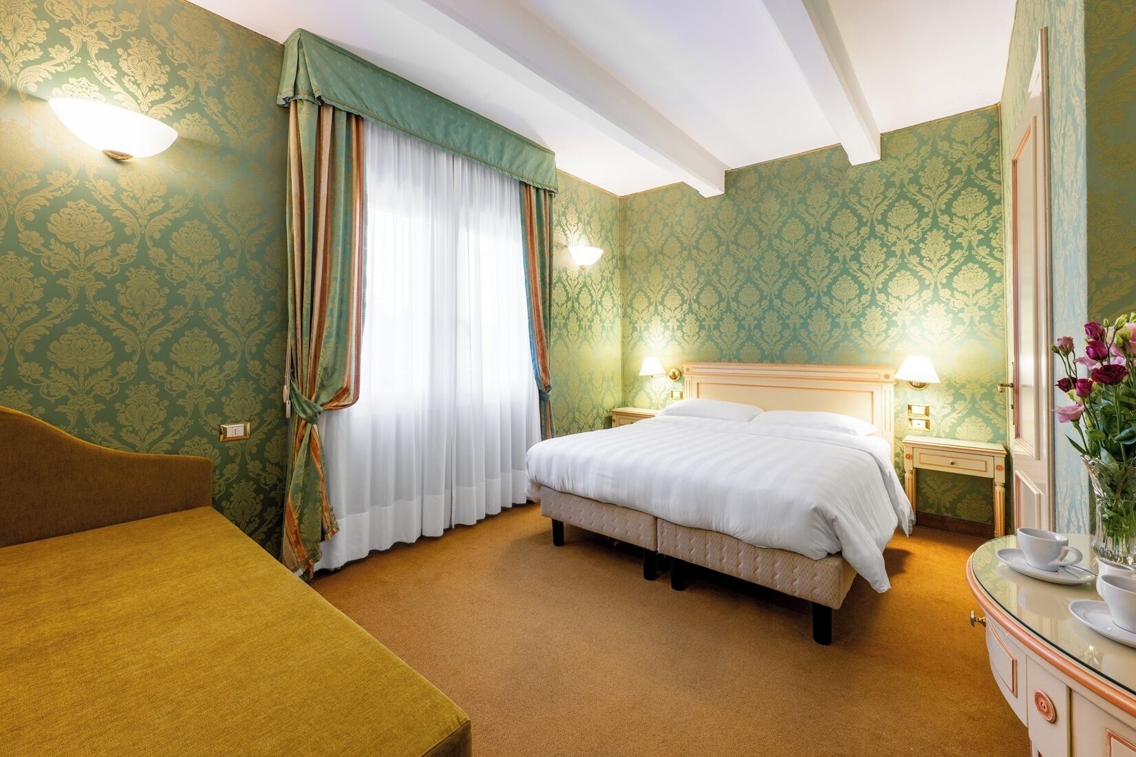 A hotel room with a bed, a bedside table with a lamp, a chair, and a vase with flowers.