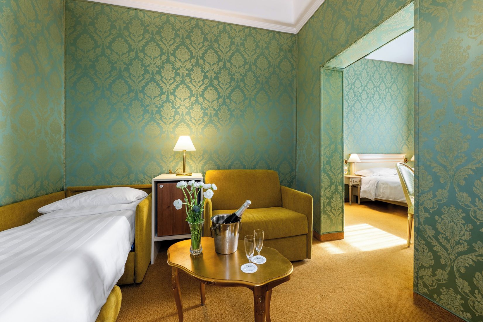 A hotel room with a bed, a sofa, a small table with a lamp, a vase with flowers, and a minibar.