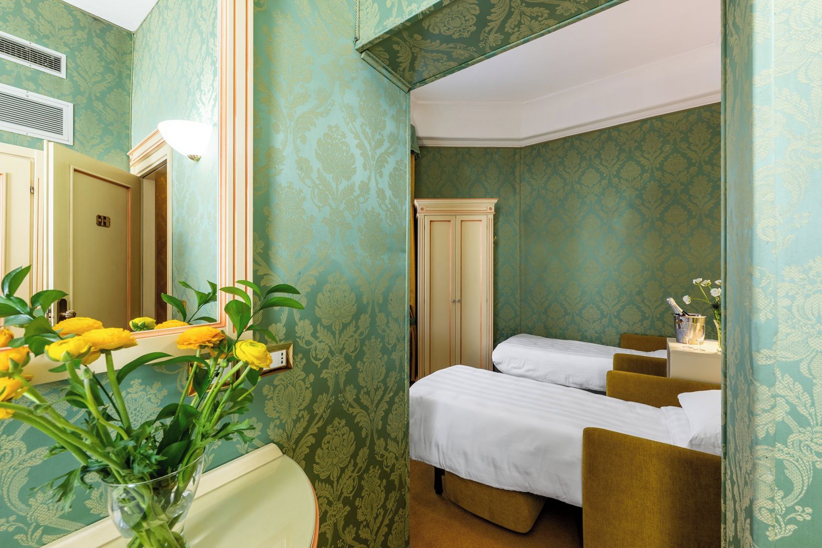 A hotel room with two beds, a mirror, a vase with flowers, and a wardrobe.