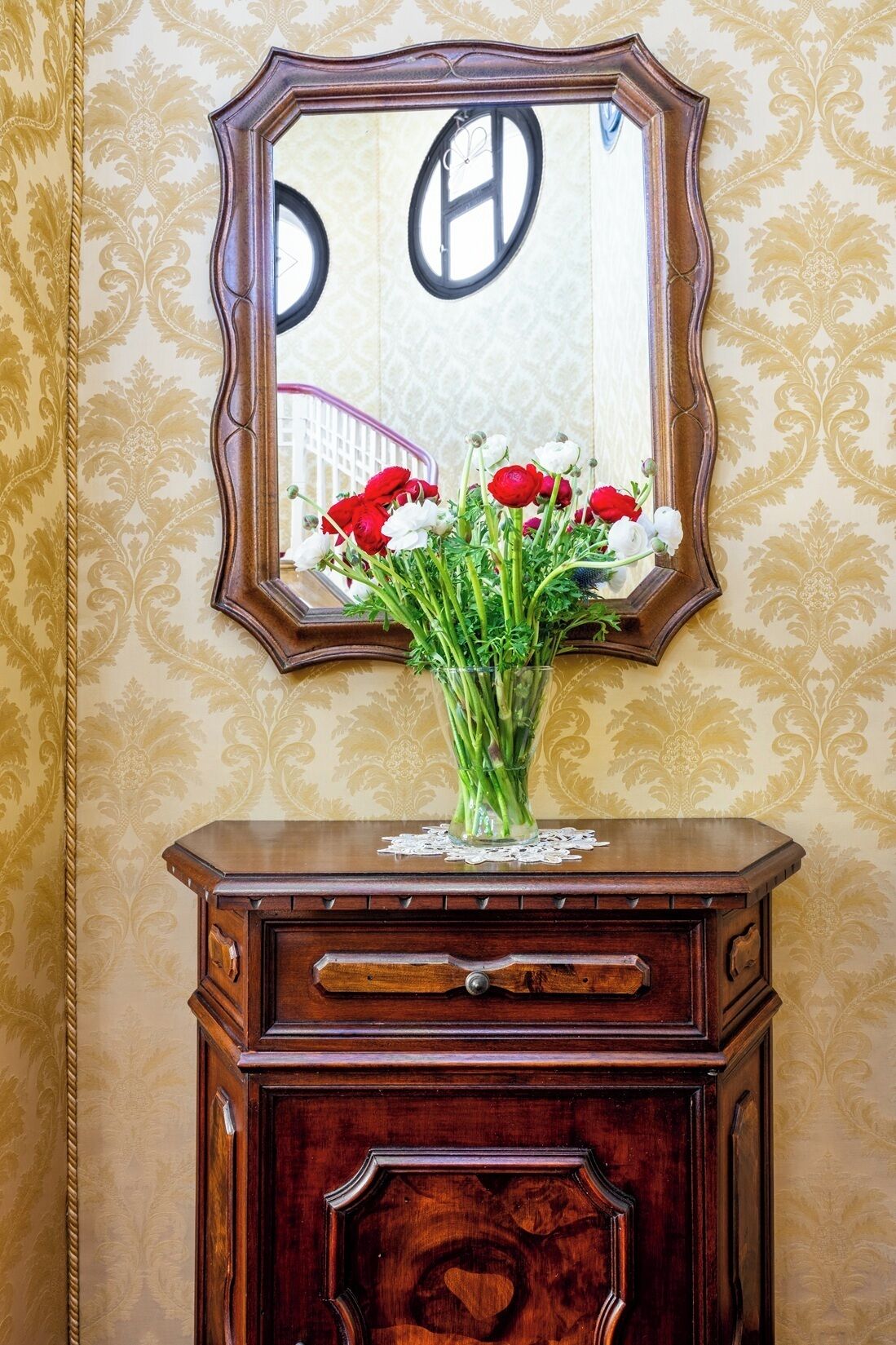 A mirror with a floral pattern, a vase with red and white flowers, and a wooden cabinet with drawers.