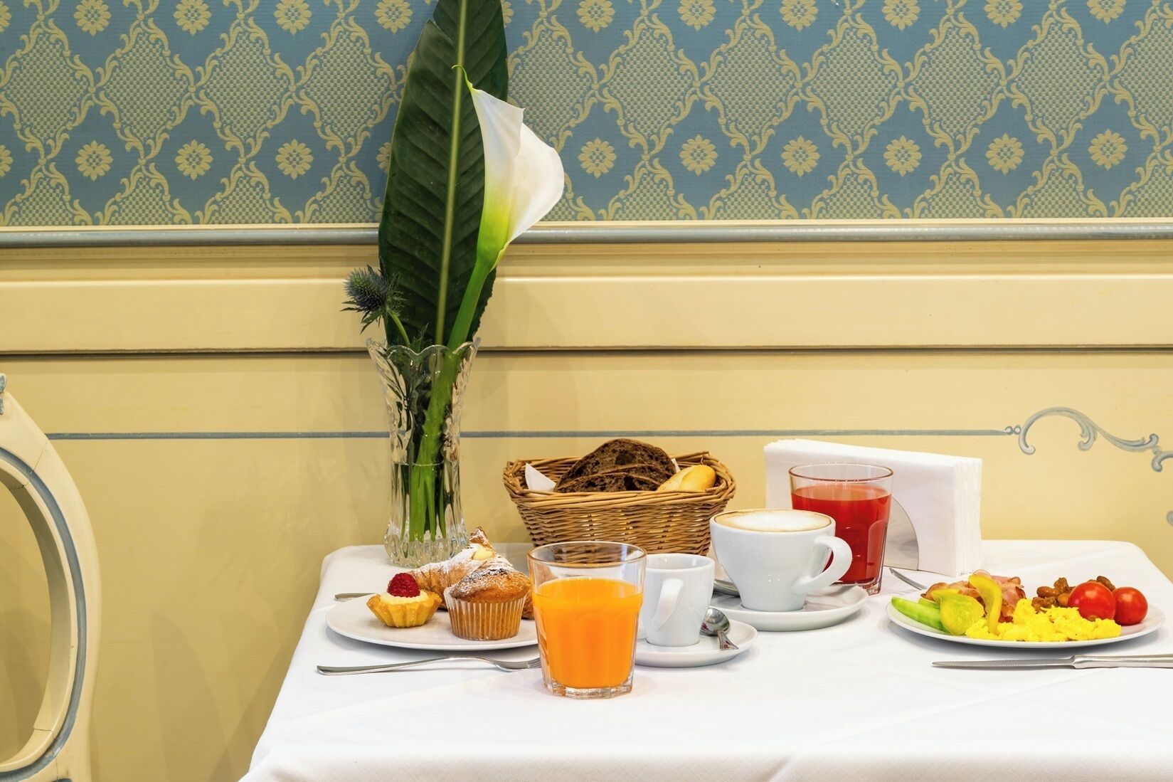 A breakfast table with a vase of flowers, a basket of bread, a plate of fruit, a glass of orange juice, and a cup of coffee.